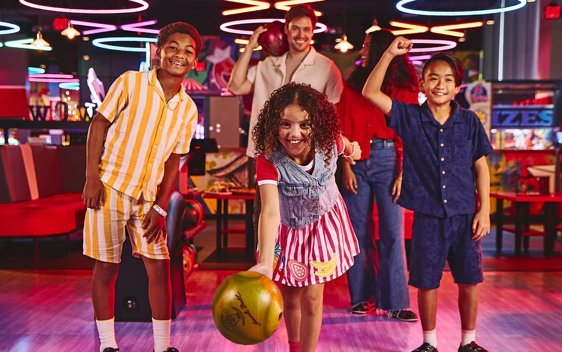 Kids and adults smiling and bowling together at a colorful bowling center, with a child rolling a bowling ball in the foreground while others cheer in a lively, neon-lit arcade and bowling environment.