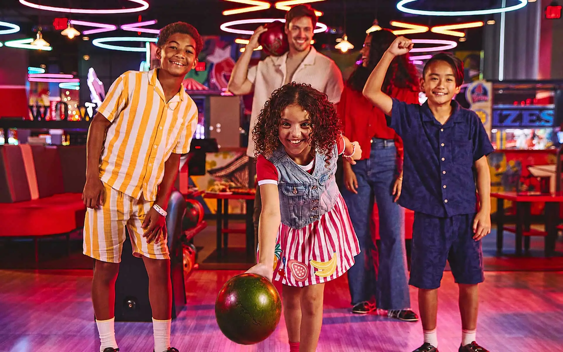 Kids and adults smiling and bowling together at a colorful bowling center, with a child rolling a bowling ball in the foreground while others cheer in a lively, neon-lit arcade and bowling environment.
