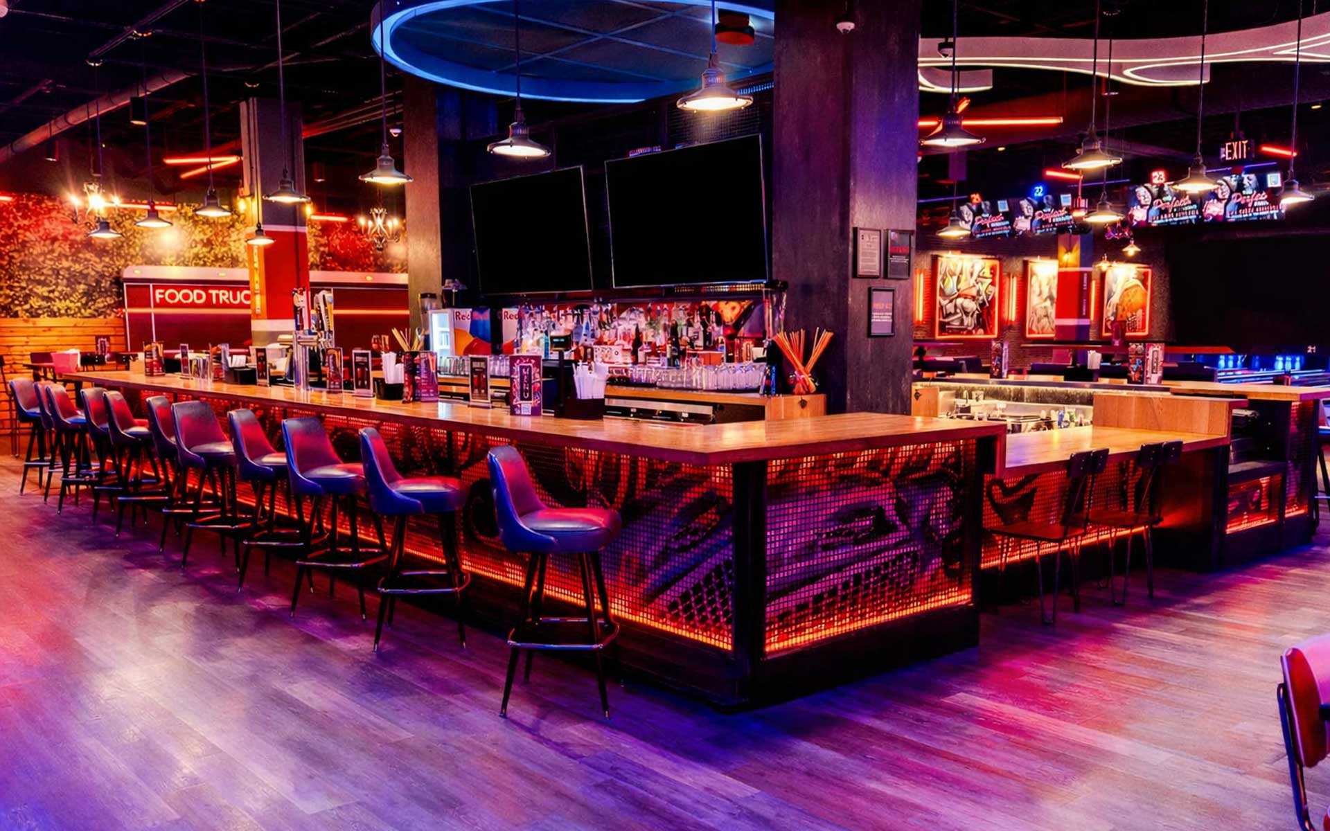 Large, modern bar inside a bowling venue featuring a glowing red-lit counter, rows of stylish bar stools, multiple TVs showing sports, and a vibrant, nightlife-inspired atmosphere