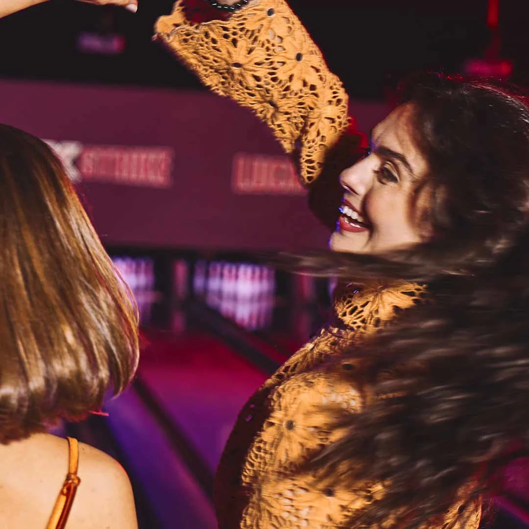 Two women dancing and laughing together at a bowling alley with neon lights in the background.