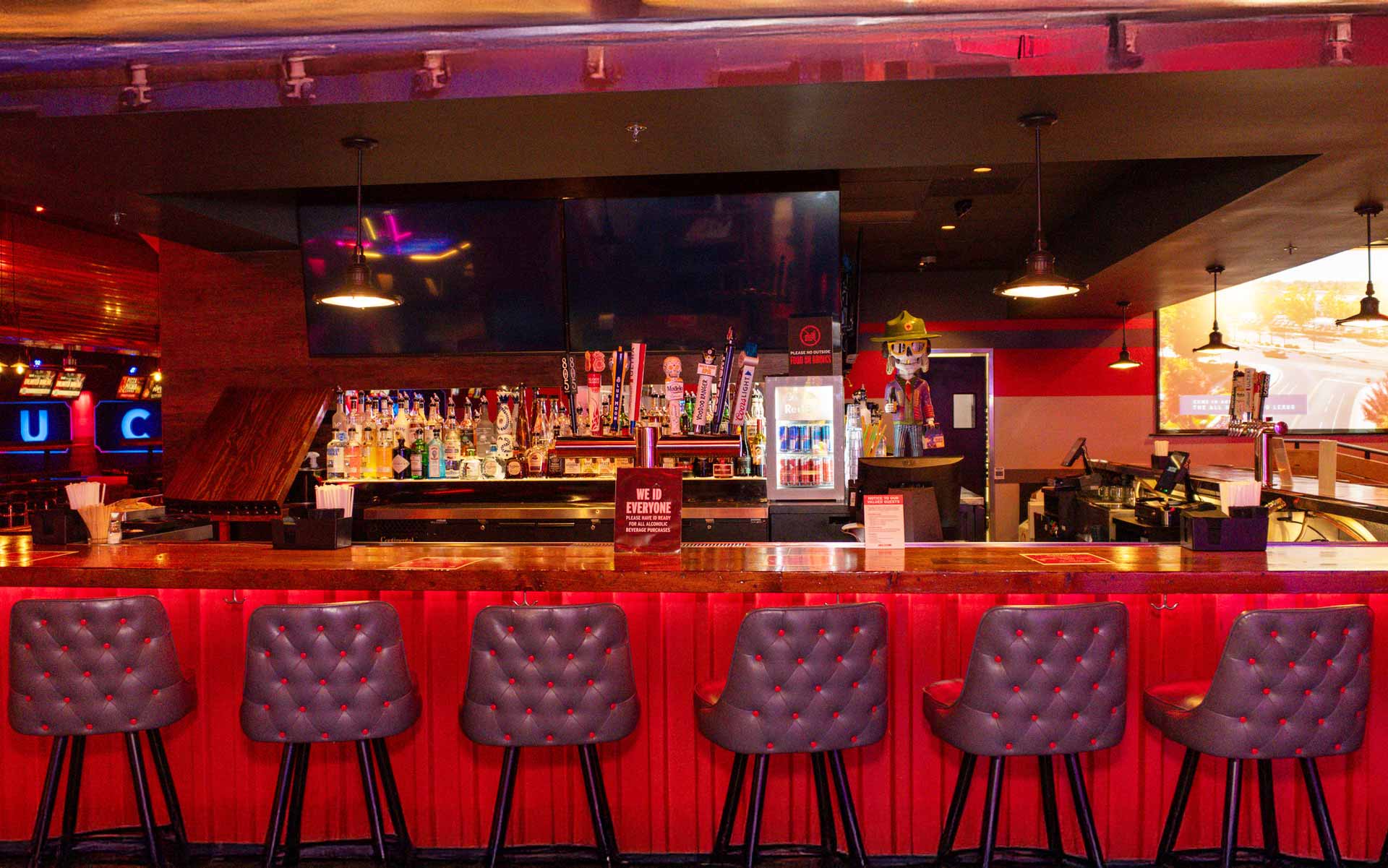 Upscale bar with a warm wood countertop, backlit red paneling, and tufted seating. Fully stocked with premium spirits, draft taps, and large-screen TVs, it serves as a central social hub with a vibrant, nightlife-inspired atmosphere.