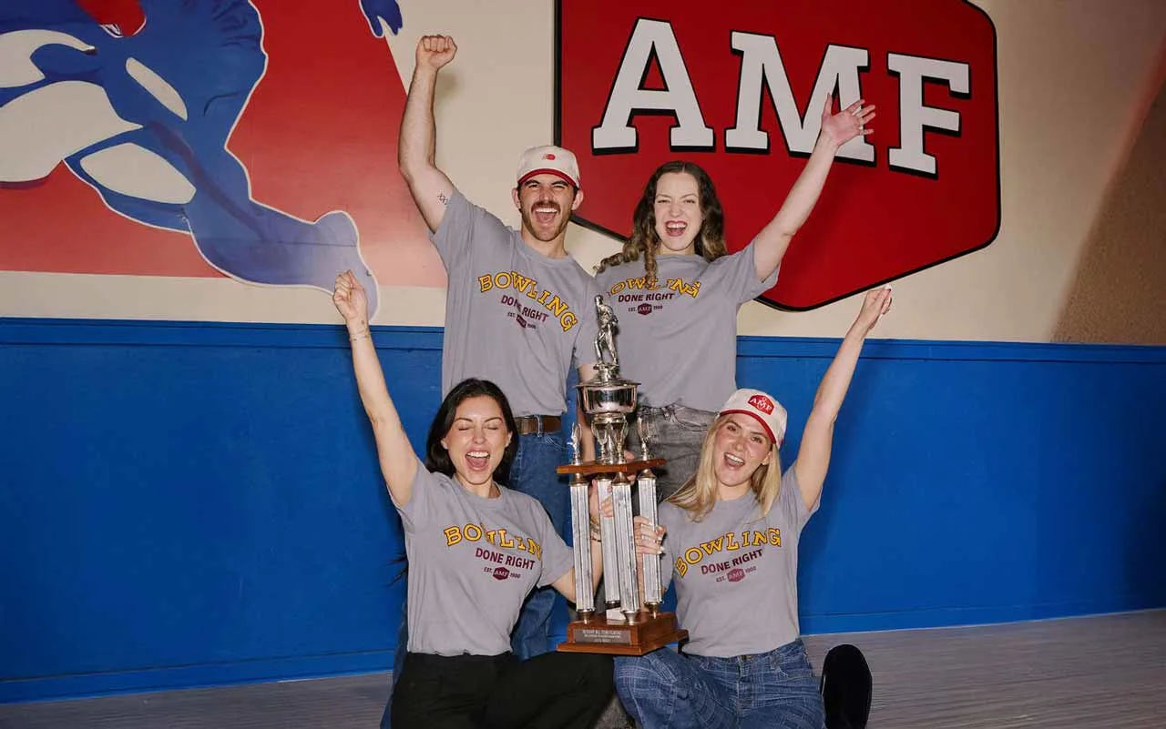 Four AMF league bowlers in "Bowling Done Right" tees cheer with a championship trophy in front of the AMF logo mural.
