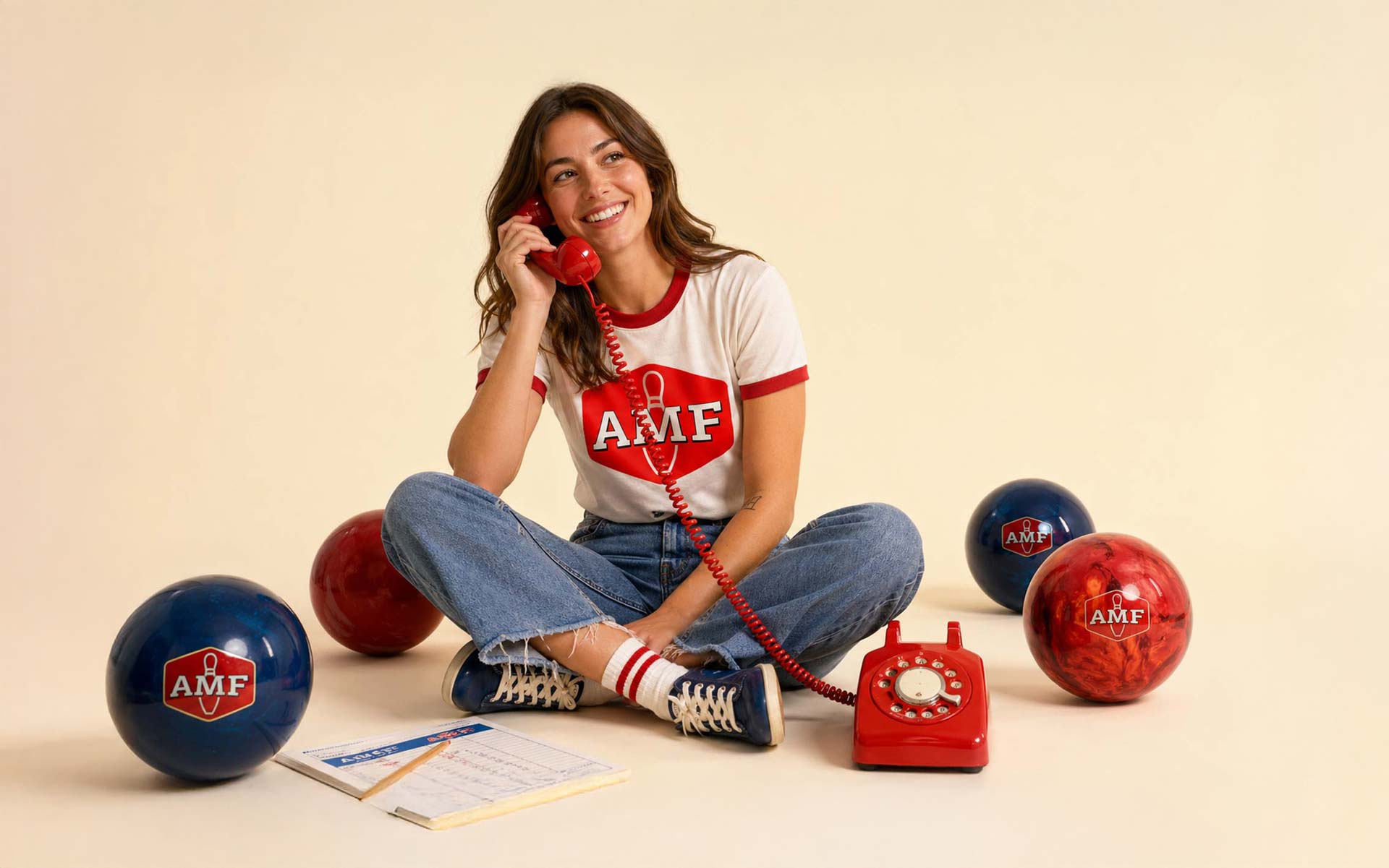 Woman in casual Americana outfit smiling on a red corded phone, seated on a clean cream backdrop with AMF bowling balls arranged around her