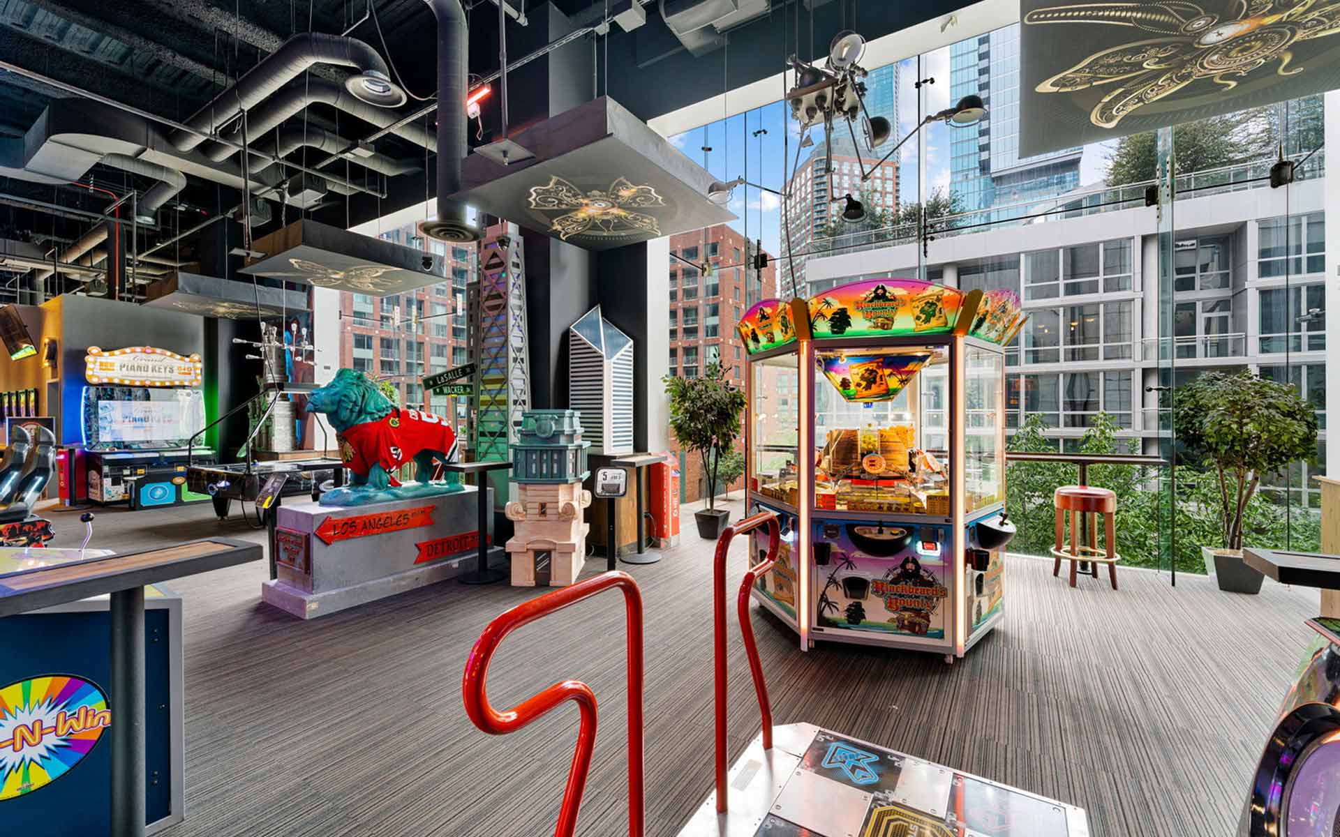 Spacious arcade with floor-to-ceiling windows overlooking a city skyline, featuring prize machines, dance games, and modern gaming stations.