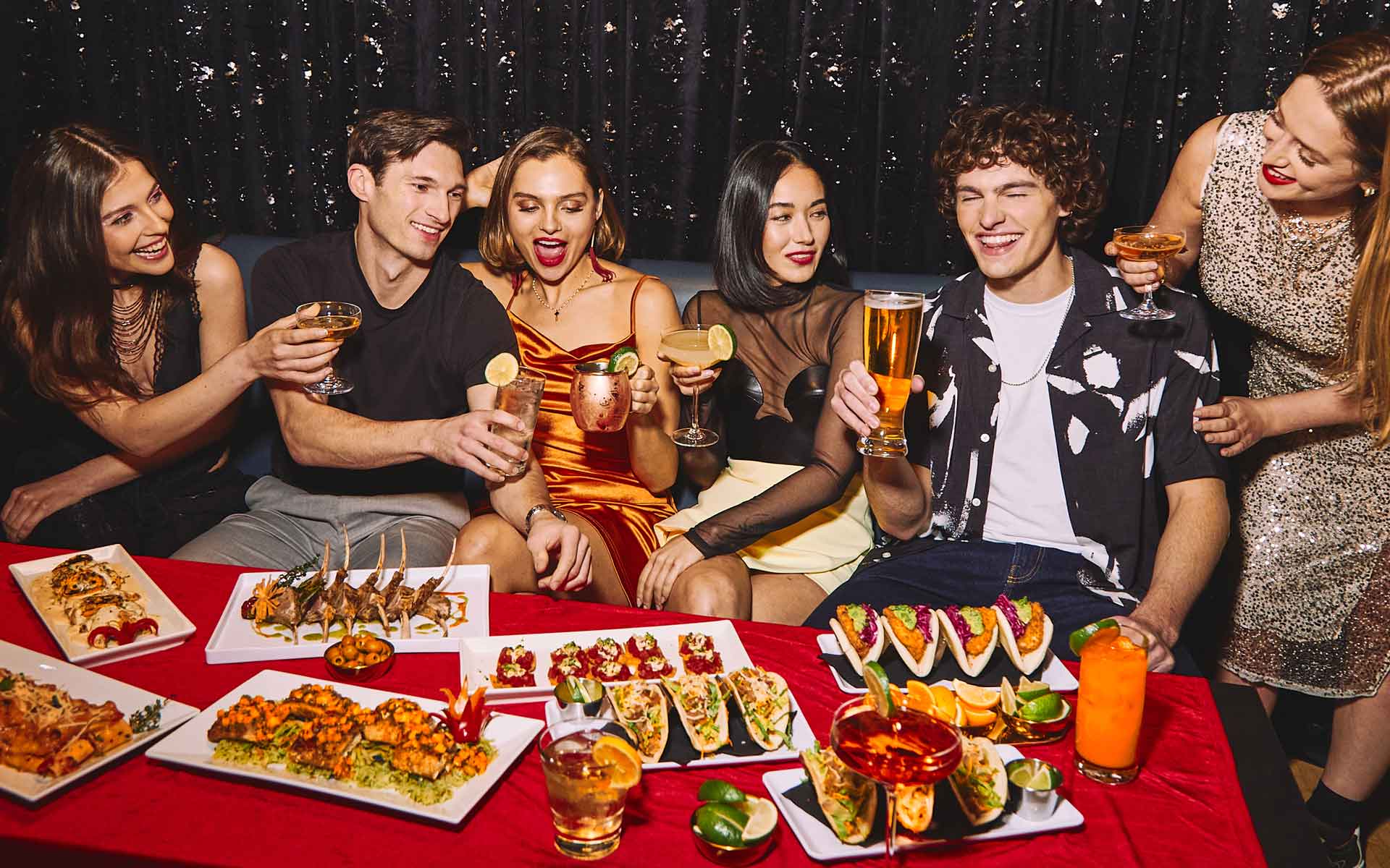 Group of friends enjoying an upscale party with craft cocktails, beer, and shareable appetizers at a Lucky Strike venue, celebrating together with food and drinks in a lively nightlife setting.