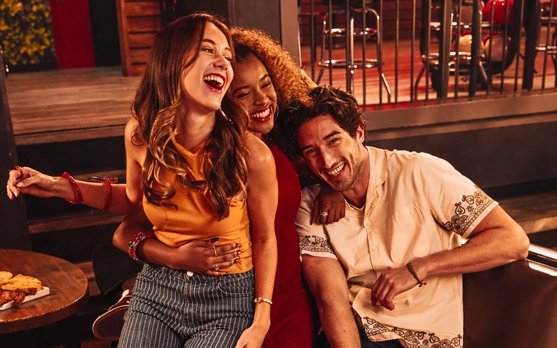 Three friends laughing together in a bowling lounge seating area, with drinks and food on a table nearby.