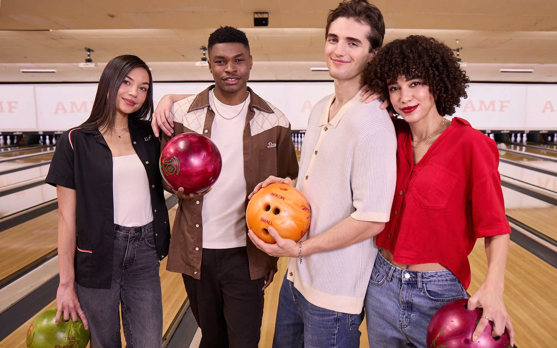 Group of four young adults smiling and holding bowling balls on a lane at an AMF bowling center, with pins and lanes visible in the background.