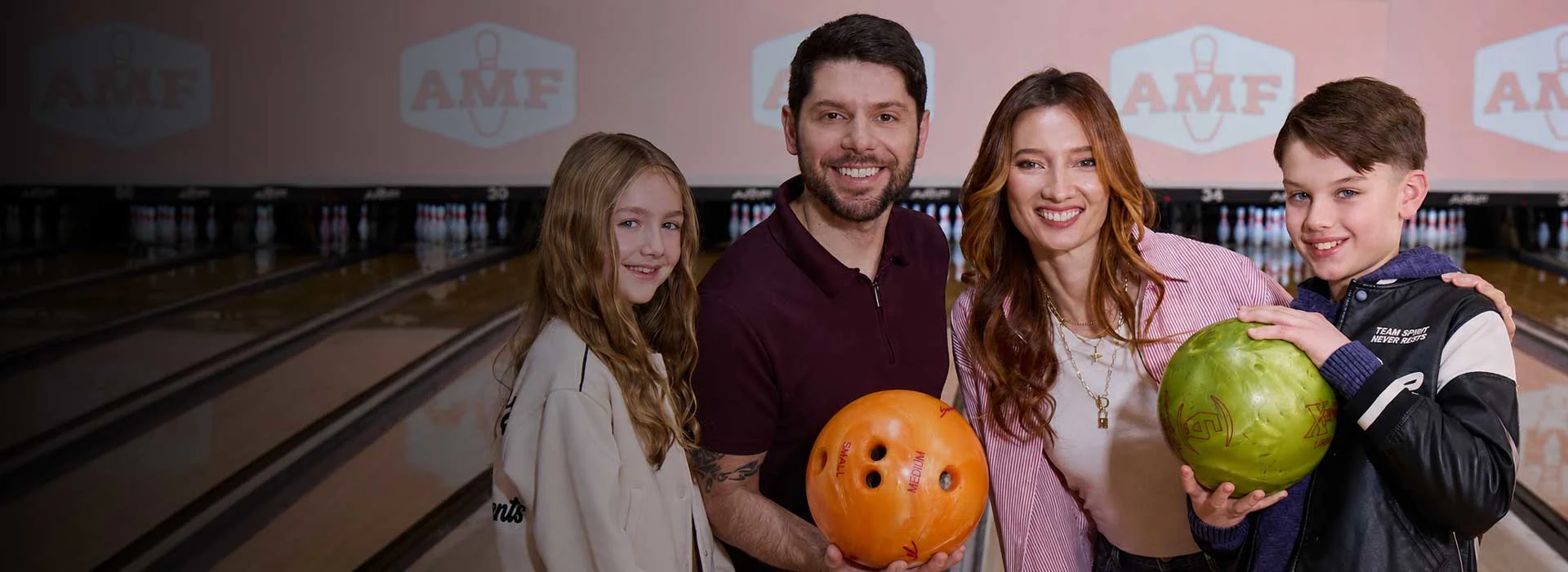 Family smiling with bowling balls at AMF, standing in front of bowling lanes with pins and AMF logos in the background.