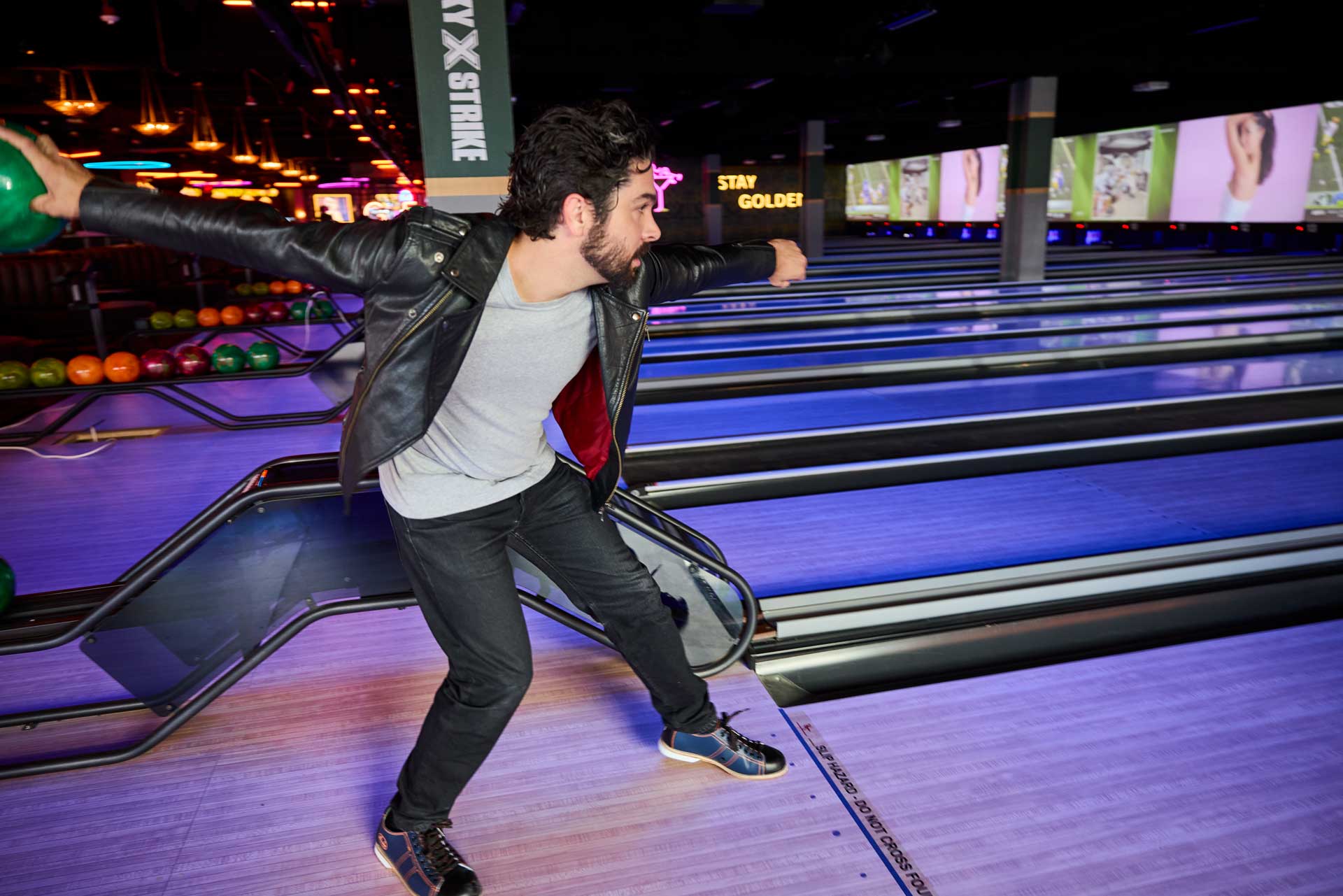 A man in a black leather jacket bowls at a neon-lit Lucky Strike bowling alley, mid-throw on glowing blue lanes with colorful TVs and ambient nightlife lighting in the background.