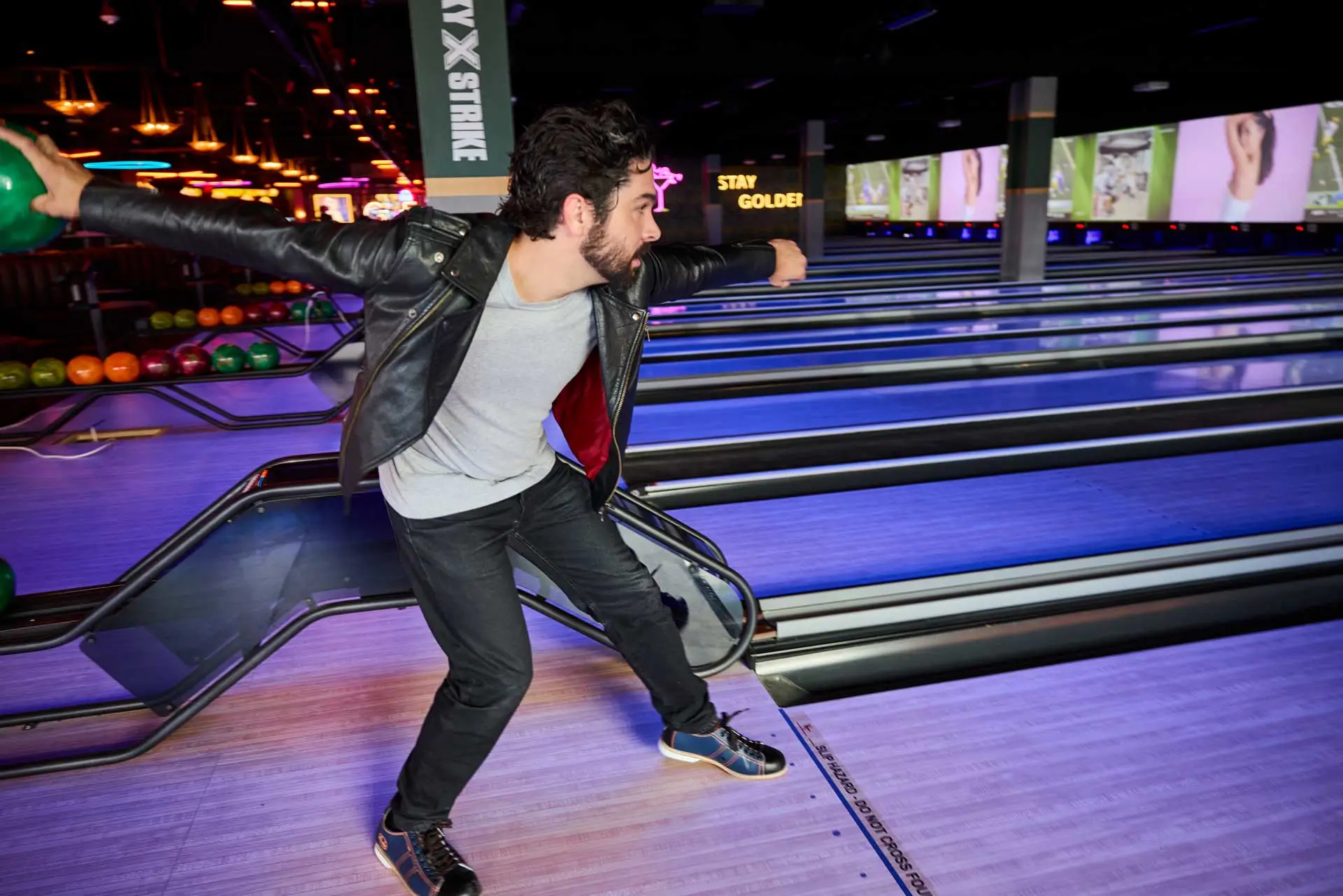 A man in a black leather jacket bowls at a neon-lit Lucky Strike bowling alley, mid-throw on glowing blue lanes with colorful TVs and ambient nightlife lighting in the background.