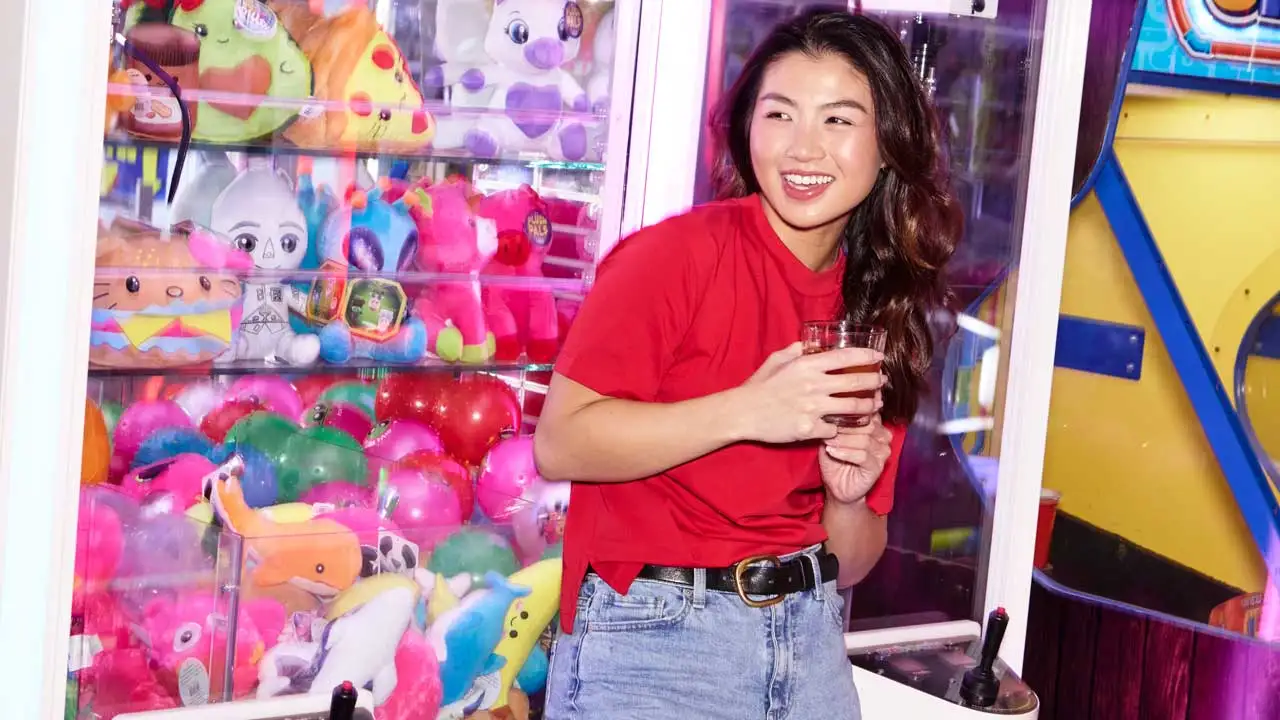 A woman holding a drink while smiling and standing in front of a crane machine.