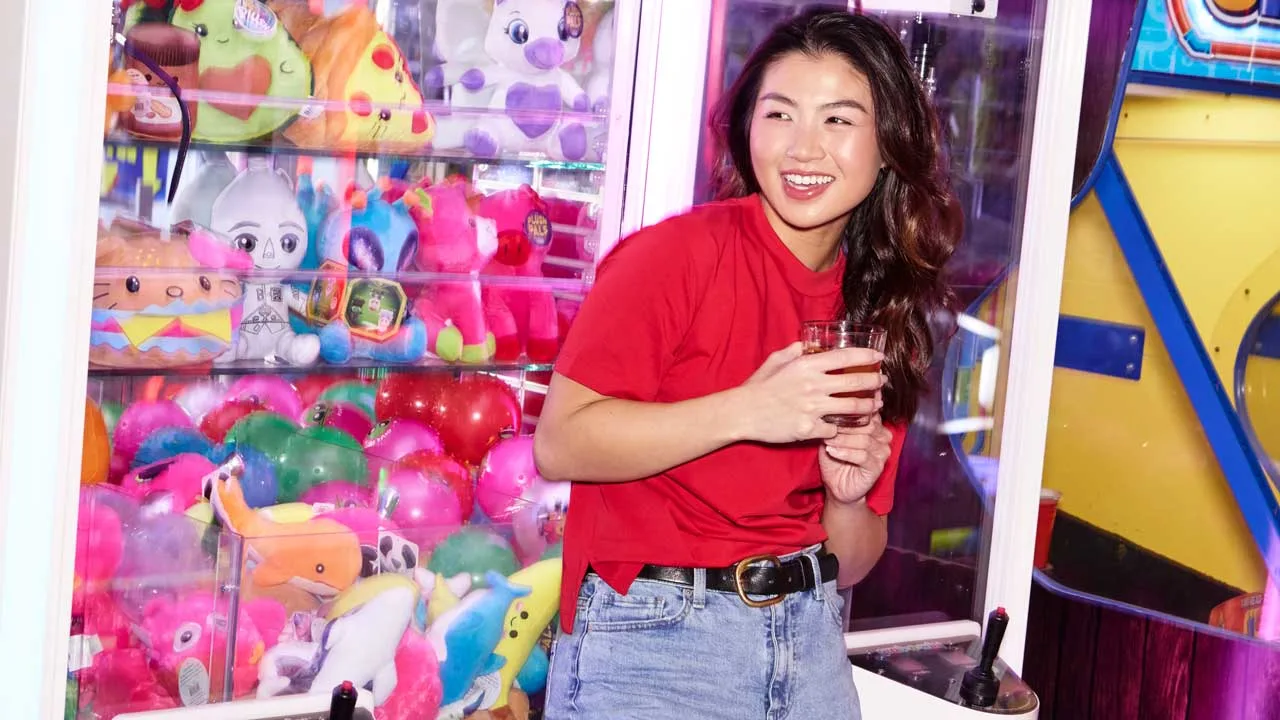 A woman holding a drink while smiling and standing in front of a crane machine.