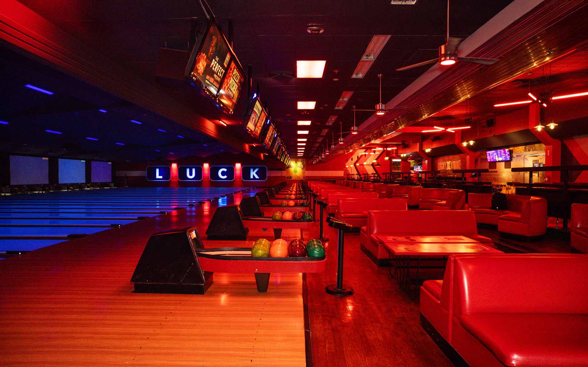 Modern bowling lanes with vibrant red and blue lighting, lounge seating, and overhead scoring screens in a sleek, nightlife-inspired setting.