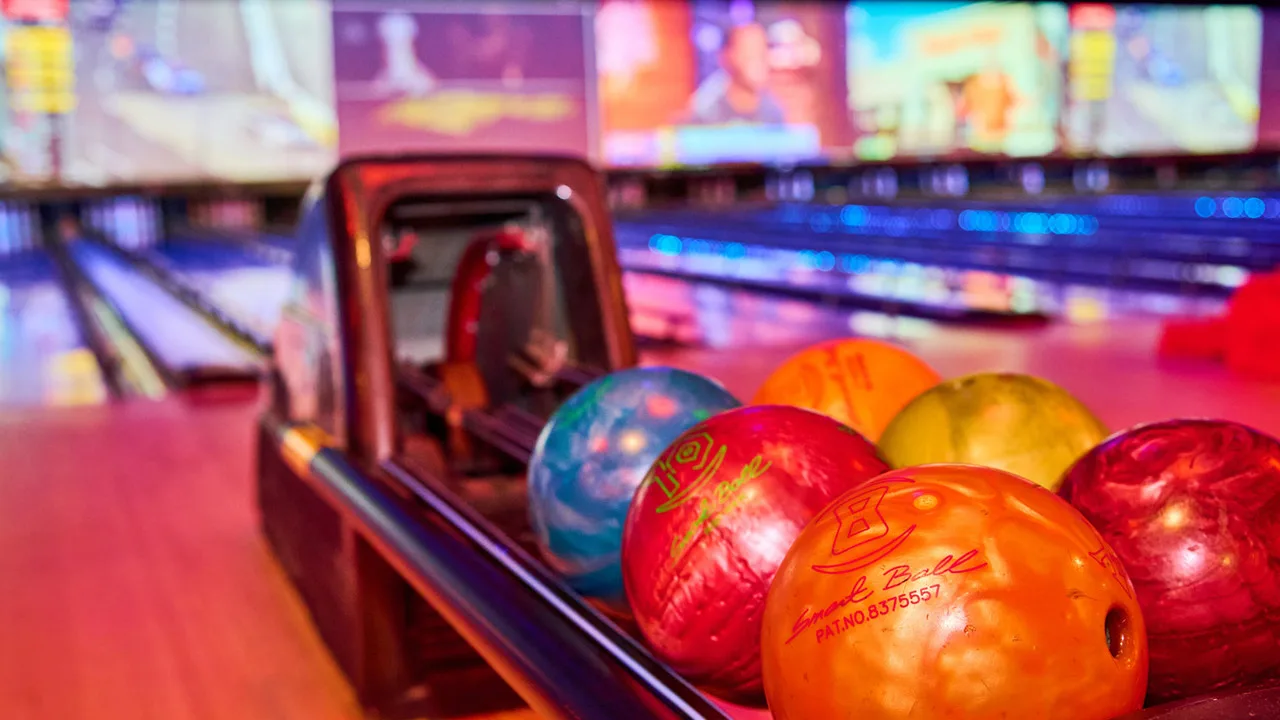 Colorful bowling balls in a return rack with bowling lanes and large TV screens in the background