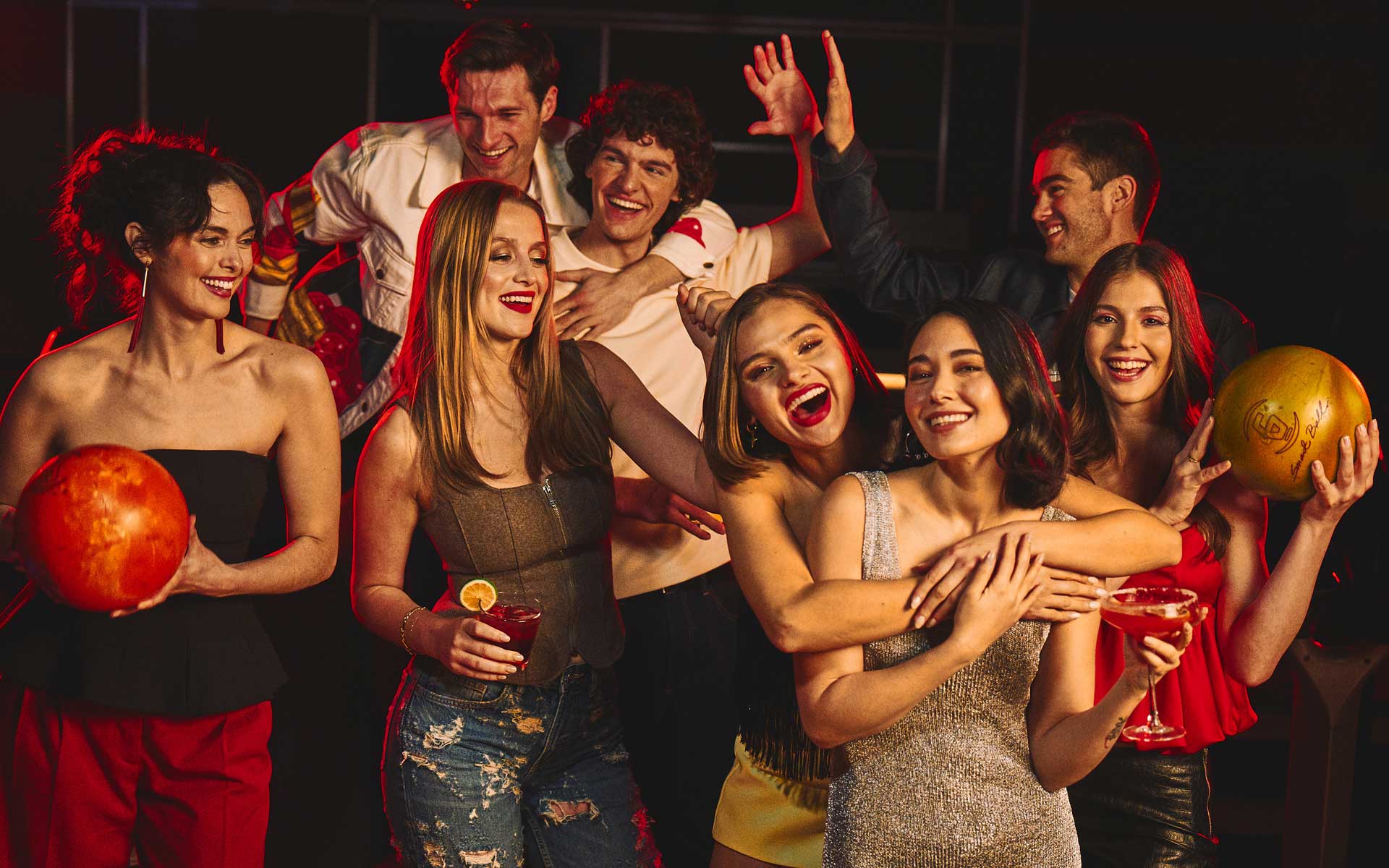 Group of friends celebrating at a bowling alley with cocktails and bowling balls, vibrant nightlife atmosphere, laughing and socializing during a night out.