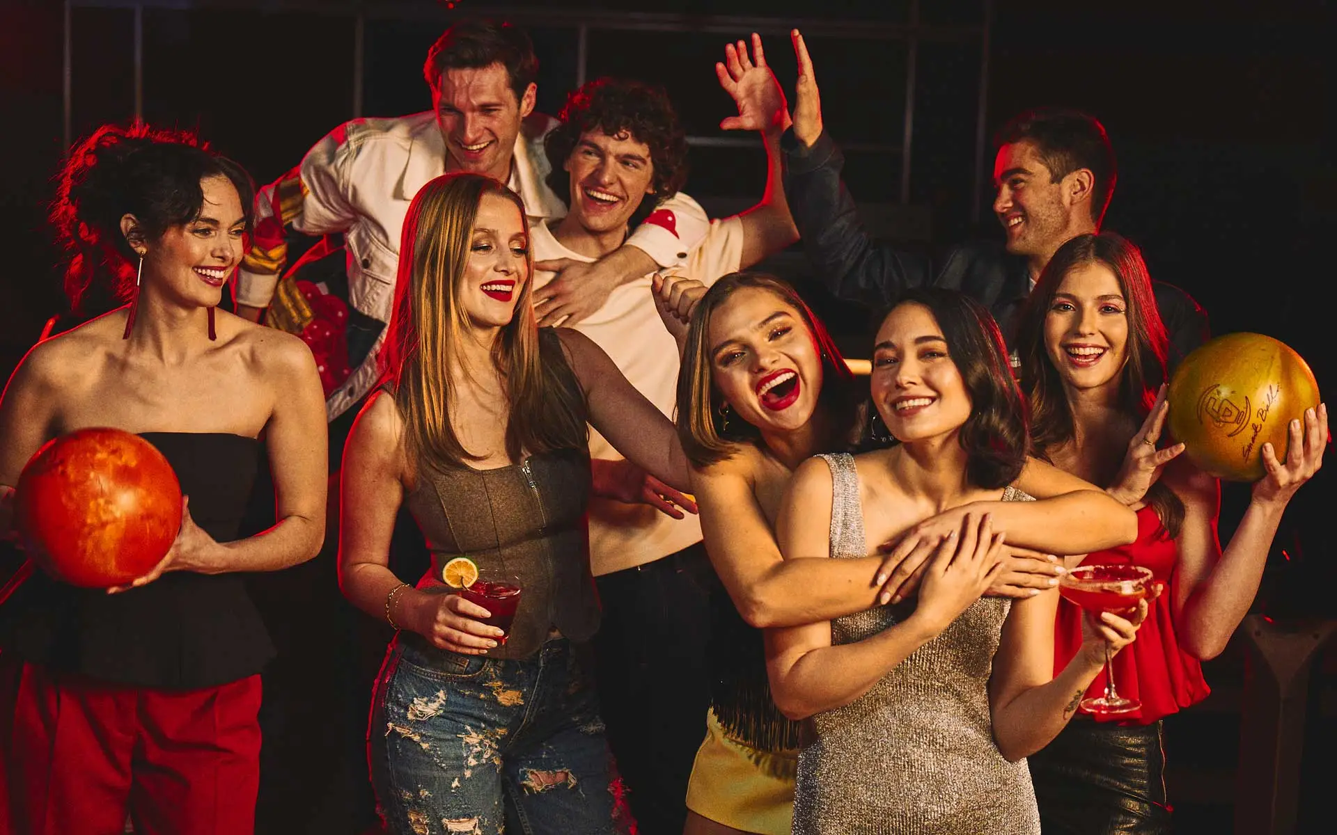 Group of friends celebrating at a bowling alley with cocktails and bowling balls, vibrant nightlife atmosphere, laughing and socializing during a night out.