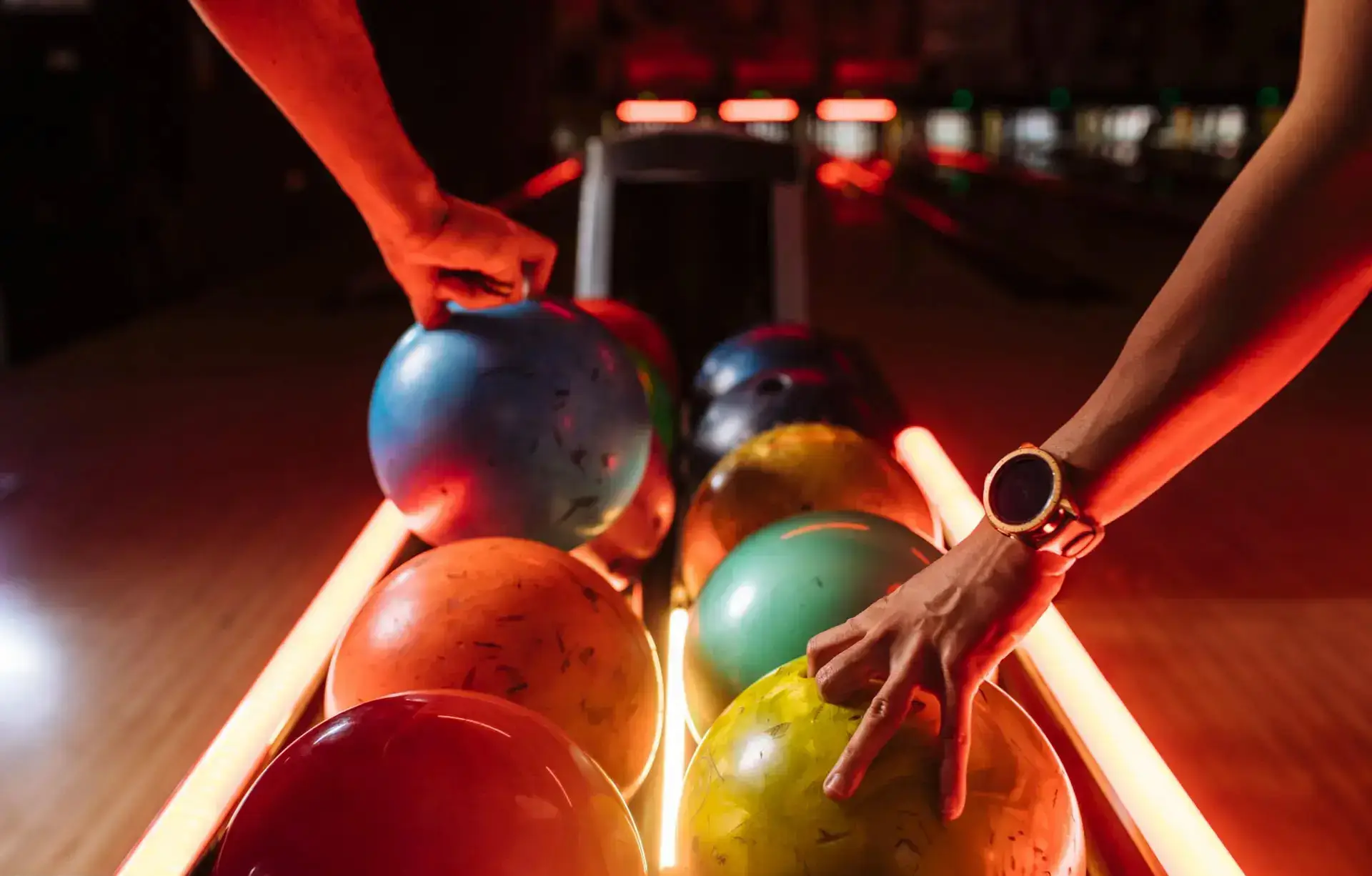 Two people reaching for bowling balls on a rack illuminated by neon lights, with glowing lanes stretching into the background.