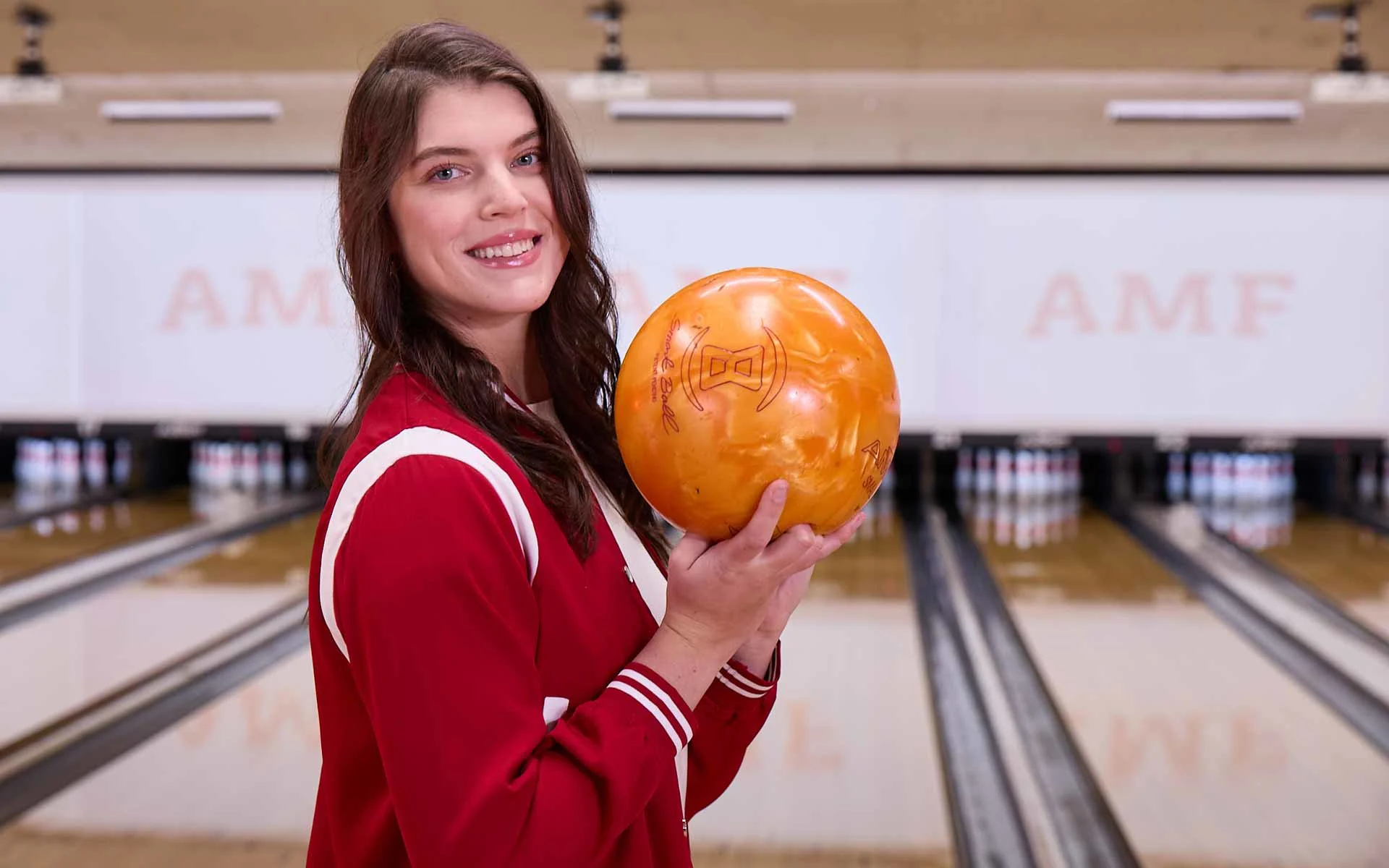Smiling woman holding an orange bowling ball at an AMF bowling alley, standing in front of the lanes with pins set in the background.