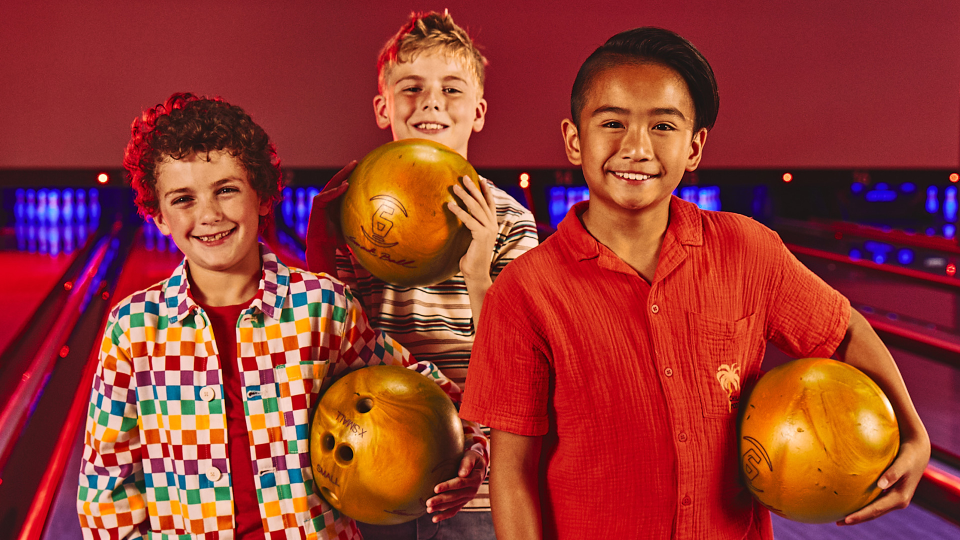 Three kids smiling and holding bowling balls on the lanes during a kids’ bowling party.