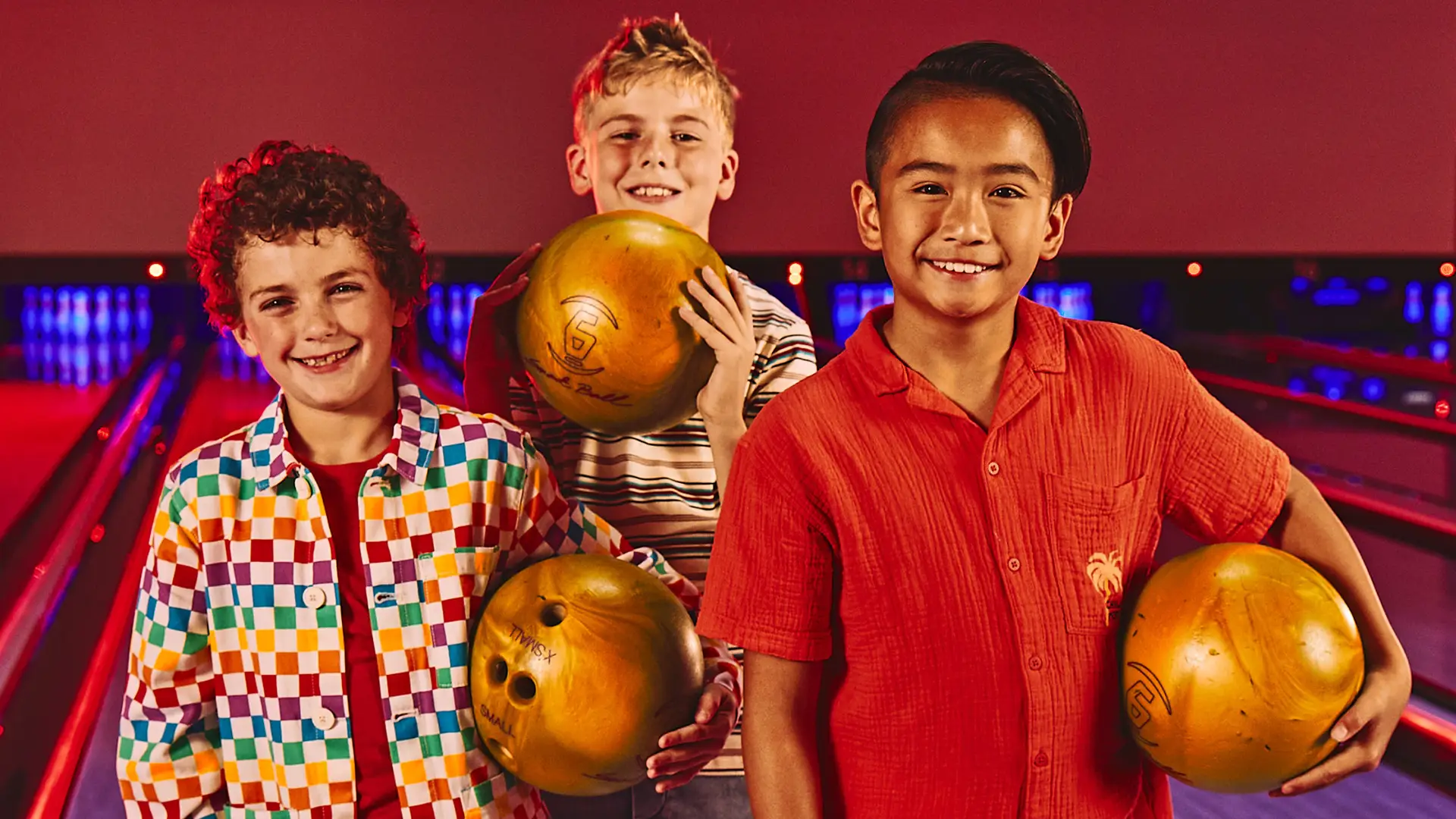 Three kids smiling and holding bowling balls on the lanes during a kids’ bowling party.