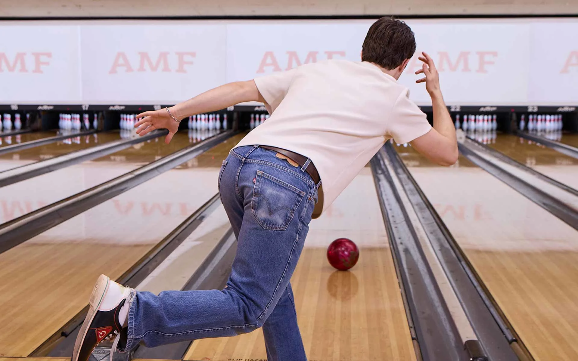 Man mid-bowl releasing a red bowling ball down the lane, captured from behind in an AMF bowling alley with pins set ahead.