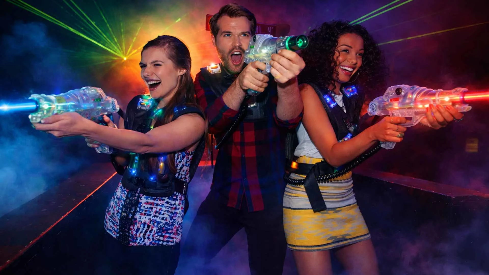 Three adults in a neon-lit laser tag arena aim glowing laser blasters through thick colorful smoke and bright green beams, creating an intense, high-energy gaming atmosphere.
