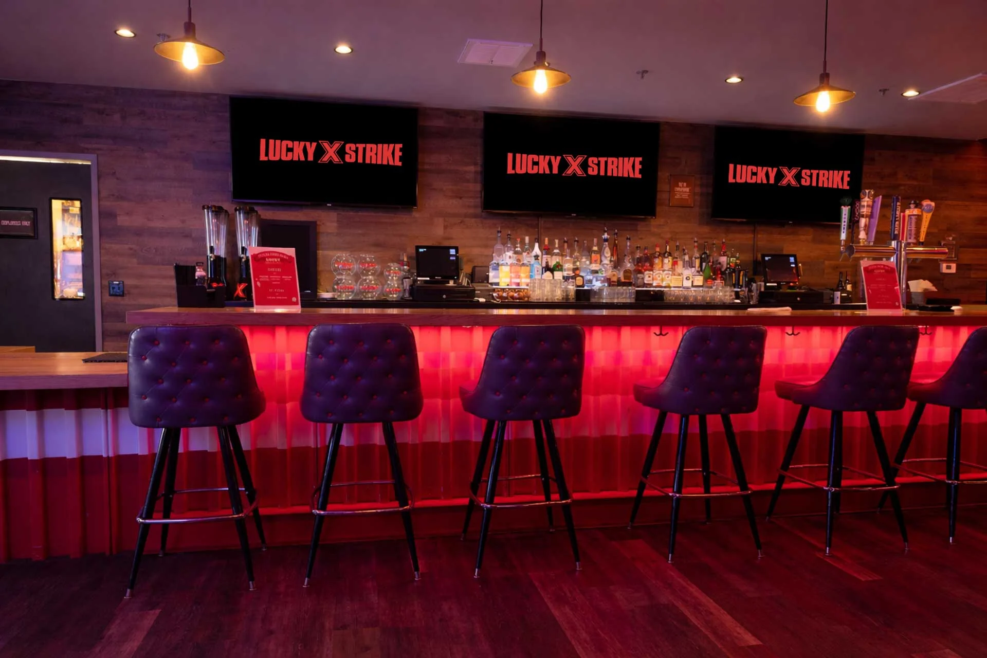 Neon-lit bar with a glowing red underlight, tufted bar stools, and three overhead screens displaying the Lucky Strike brand above a fully stocked back bar.