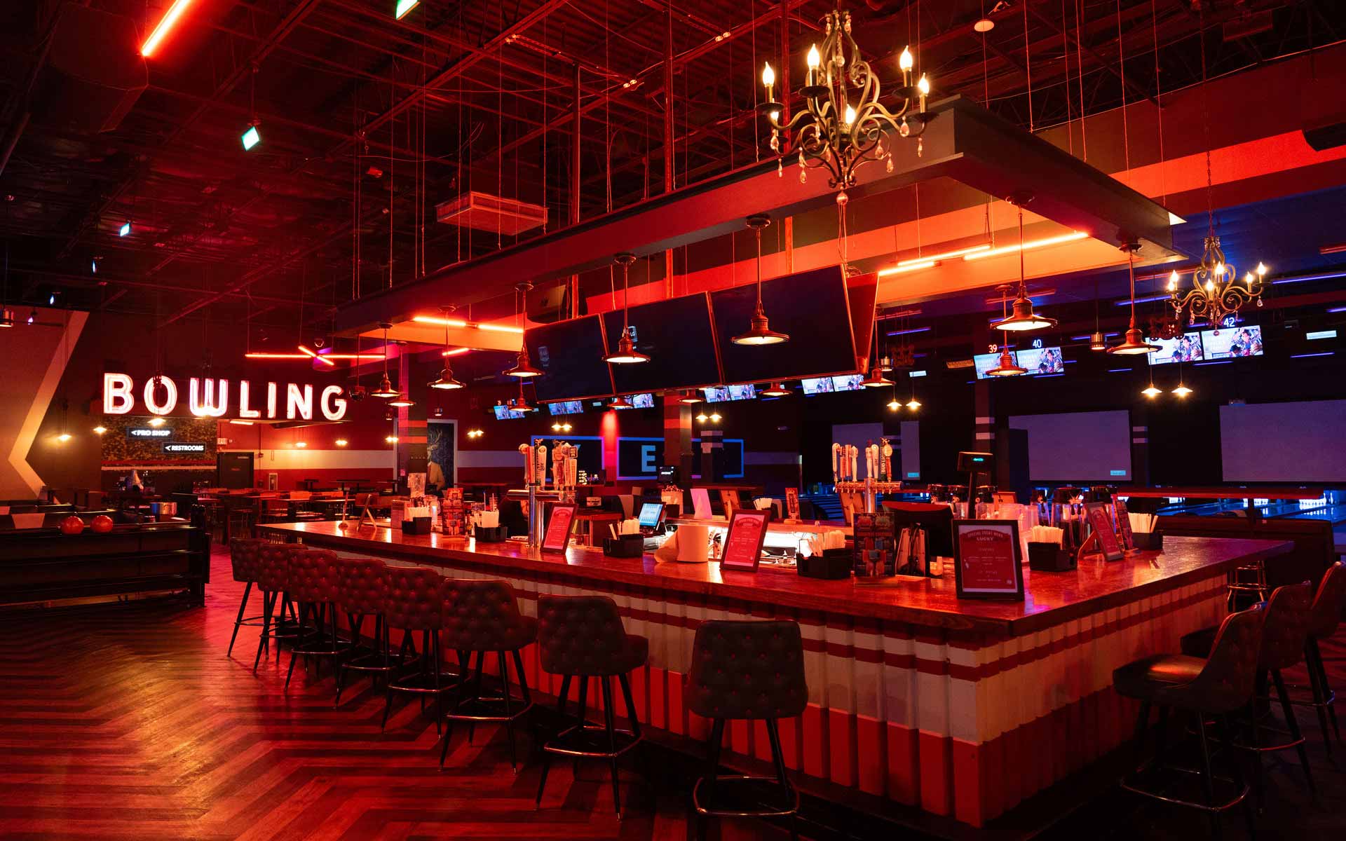 Upscale Lucky Strike bar with red ambient lighting, bowling lanes in the background, and a large central bar with seating and draft taps.