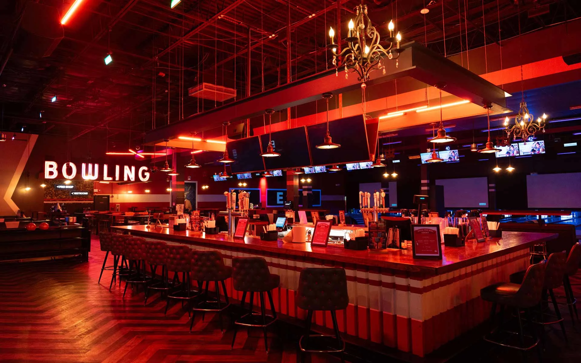 Upscale Lucky Strike bar with red ambient lighting, bowling lanes in the background, and a large central bar with seating and draft taps.