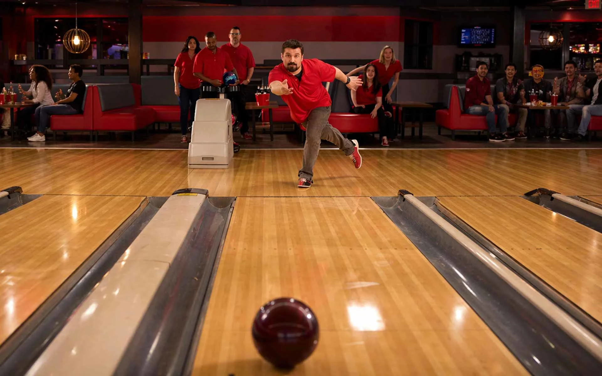Man mid-bowl releasing a ball down a polished lane, with ball returns on both sides and groups of friends socializing and cheering in a modern bowling alley.
