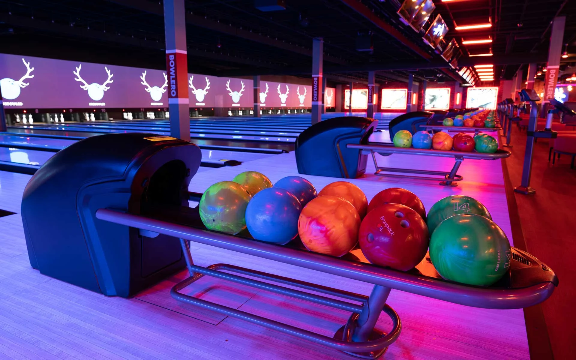 Colorful bowling balls lined up beside glowing neon-lit lanes inside a modern Bowlero bowling center at night.