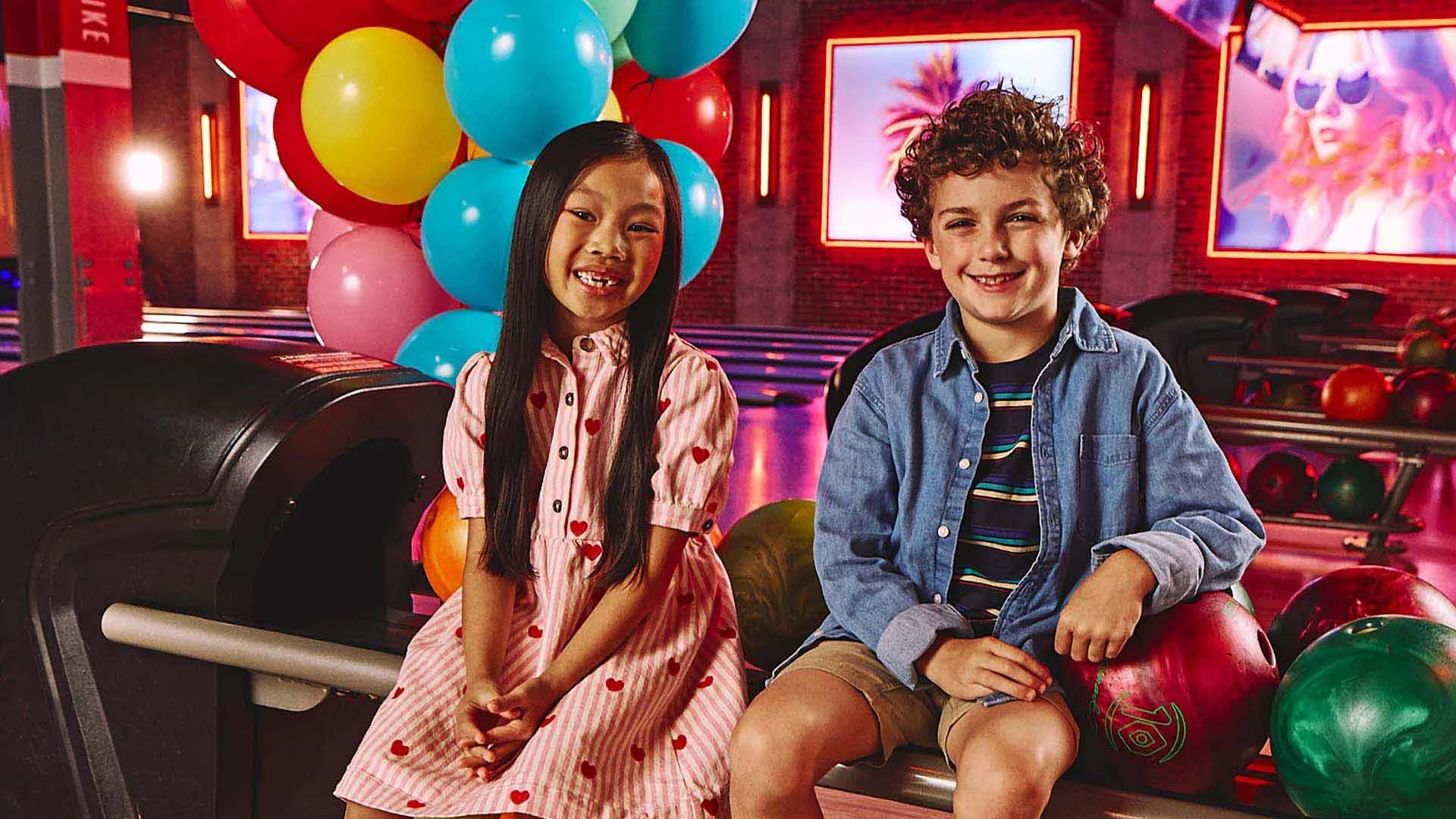 Two kids sitting by a bowling alley with party balloons in the background