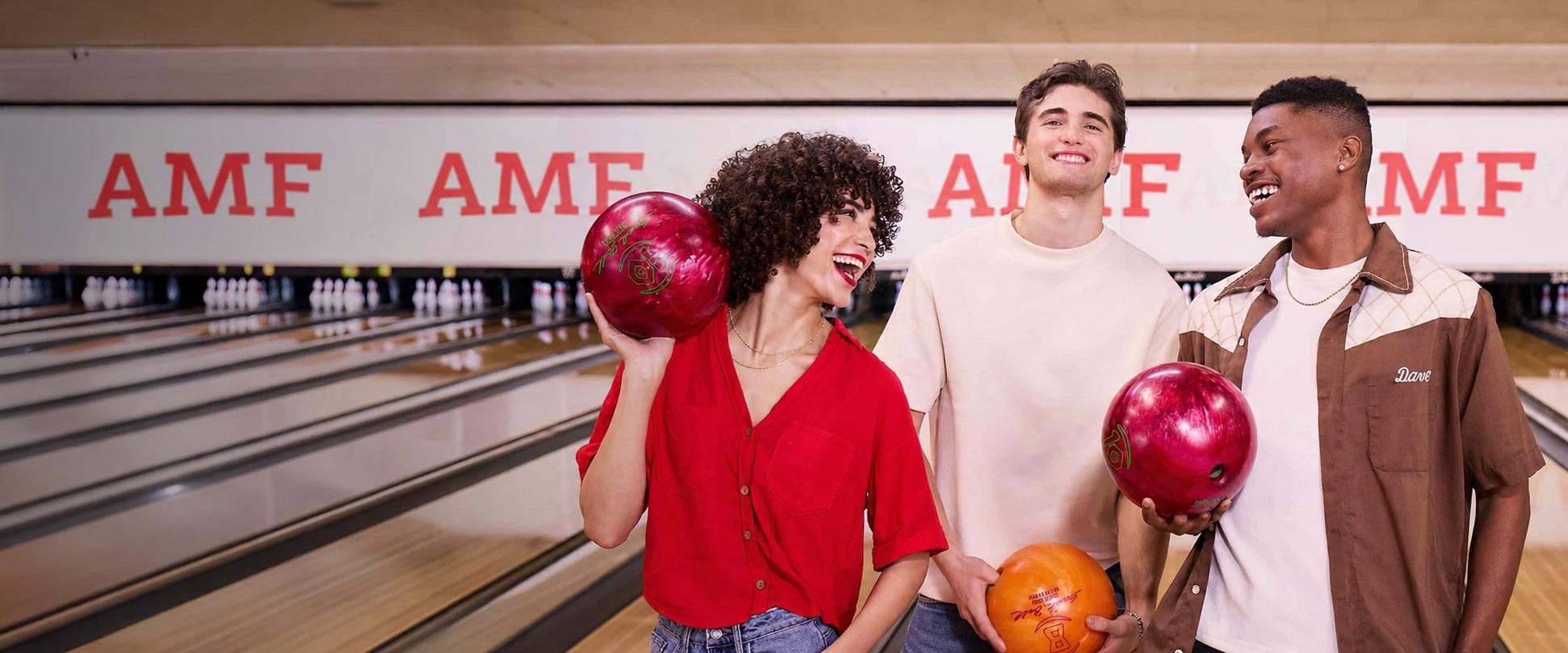 Three young adults laugh together while holding bowling balls at an AMF bowling alley, standing beside polished lanes with faint AMF logos projected across the white lane screens in the background.