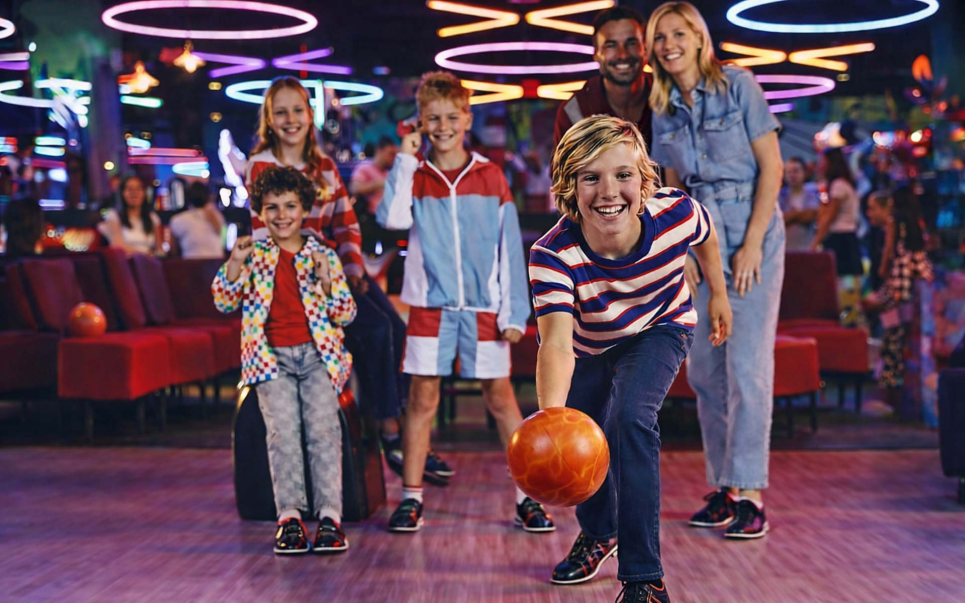 A family bowling together as a child rolls a bowling ball down the lane, with neon lights and arcade games in the background.