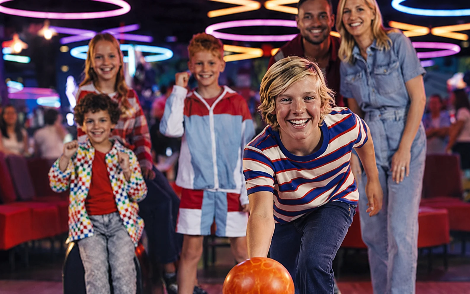 A family bowling together as a child rolls a bowling ball down the lane, with neon lights and arcade games in the background.