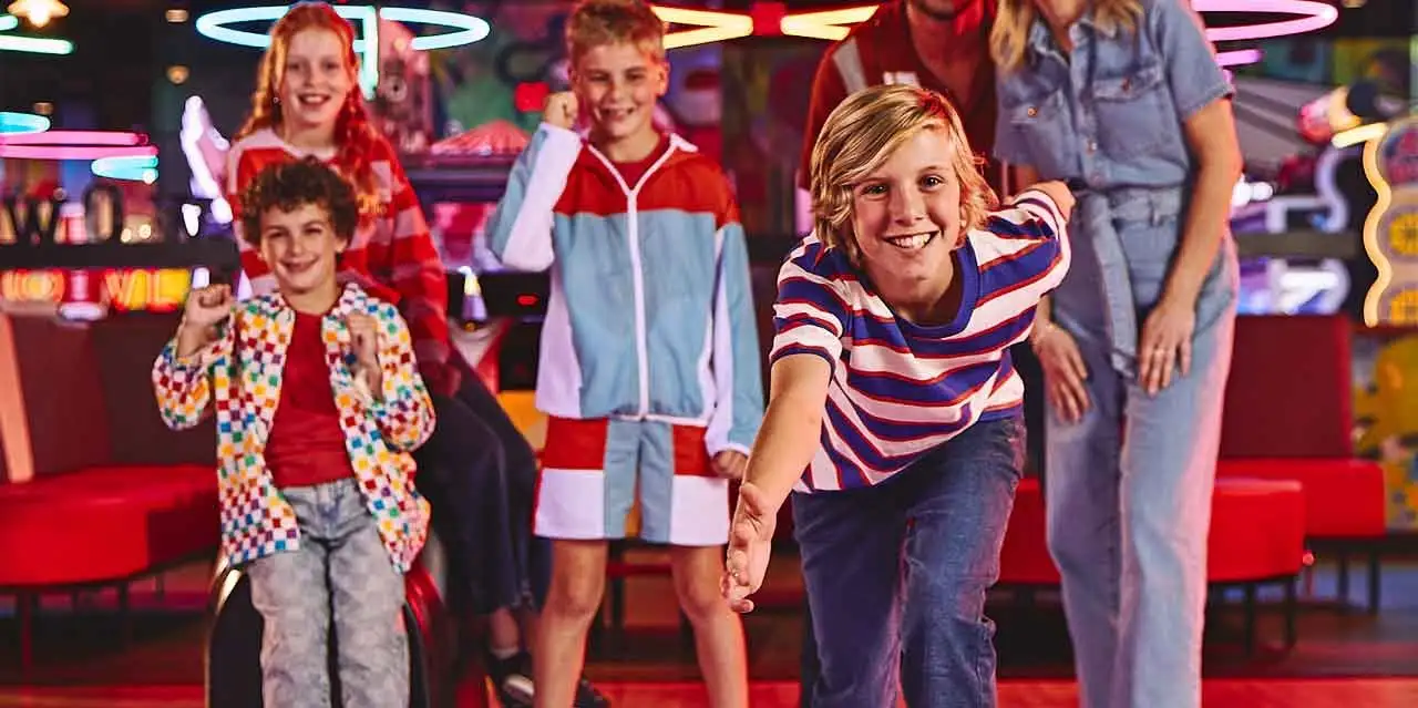 A family bowling together as a child rolls a bowling ball down the lane, with neon lights and arcade games in the background.