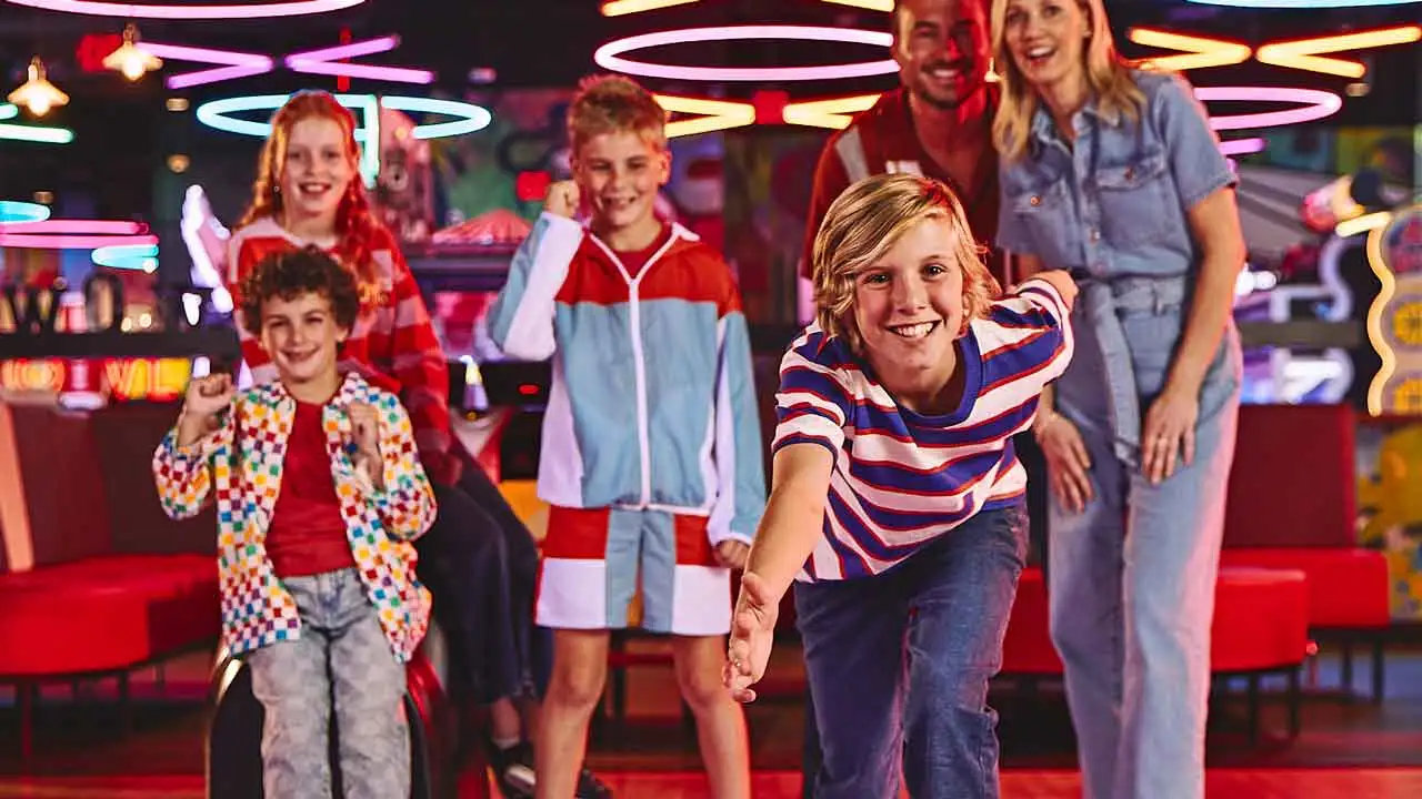 A family bowling together as a child rolls a bowling ball down the lane, with neon lights and arcade games in the background.