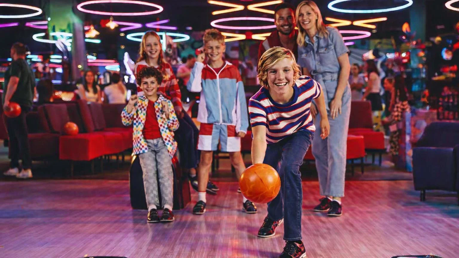 A family bowling together as a child rolls a bowling ball down the lane, with neon lights and arcade games in the background.