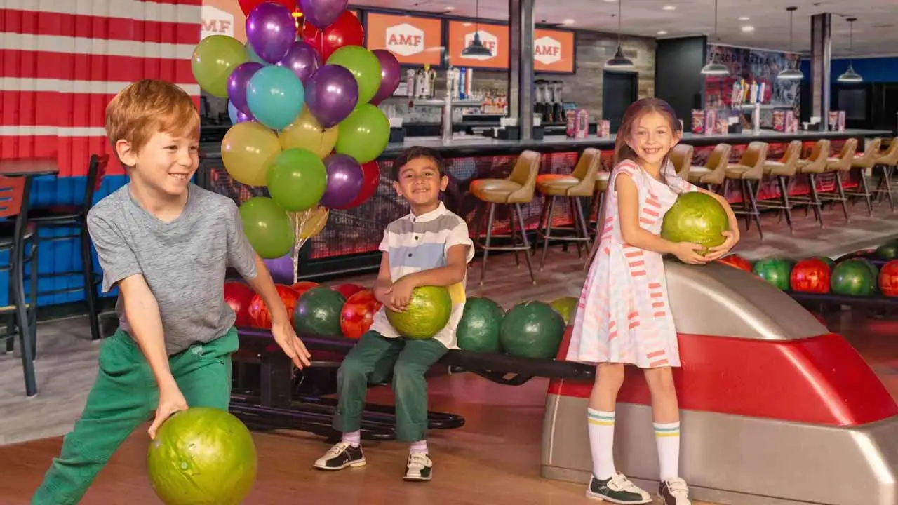 Three smiling kids having fun at an AMF bowling center, with two holding green bowling balls near a ball return while colorful balloons fill the background.