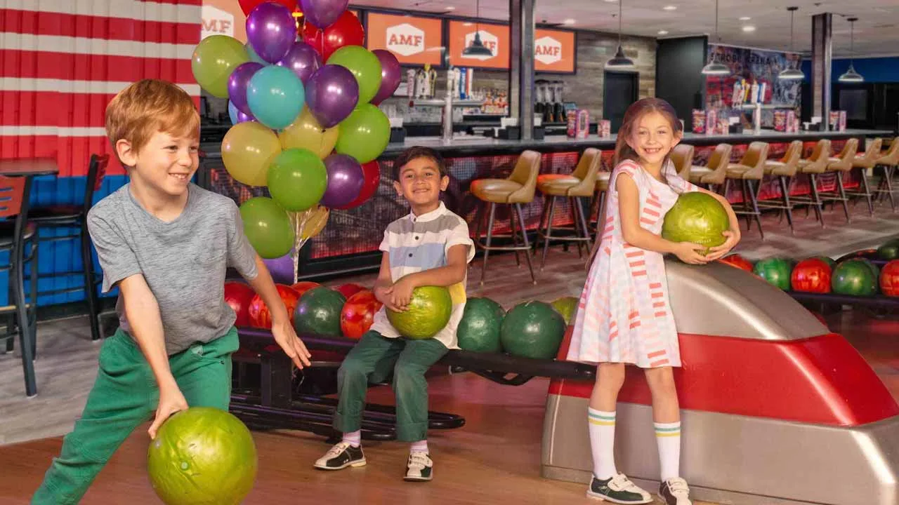 Three smiling kids having fun at an AMF bowling center, with two holding green bowling balls near a ball return while colorful balloons fill the background.