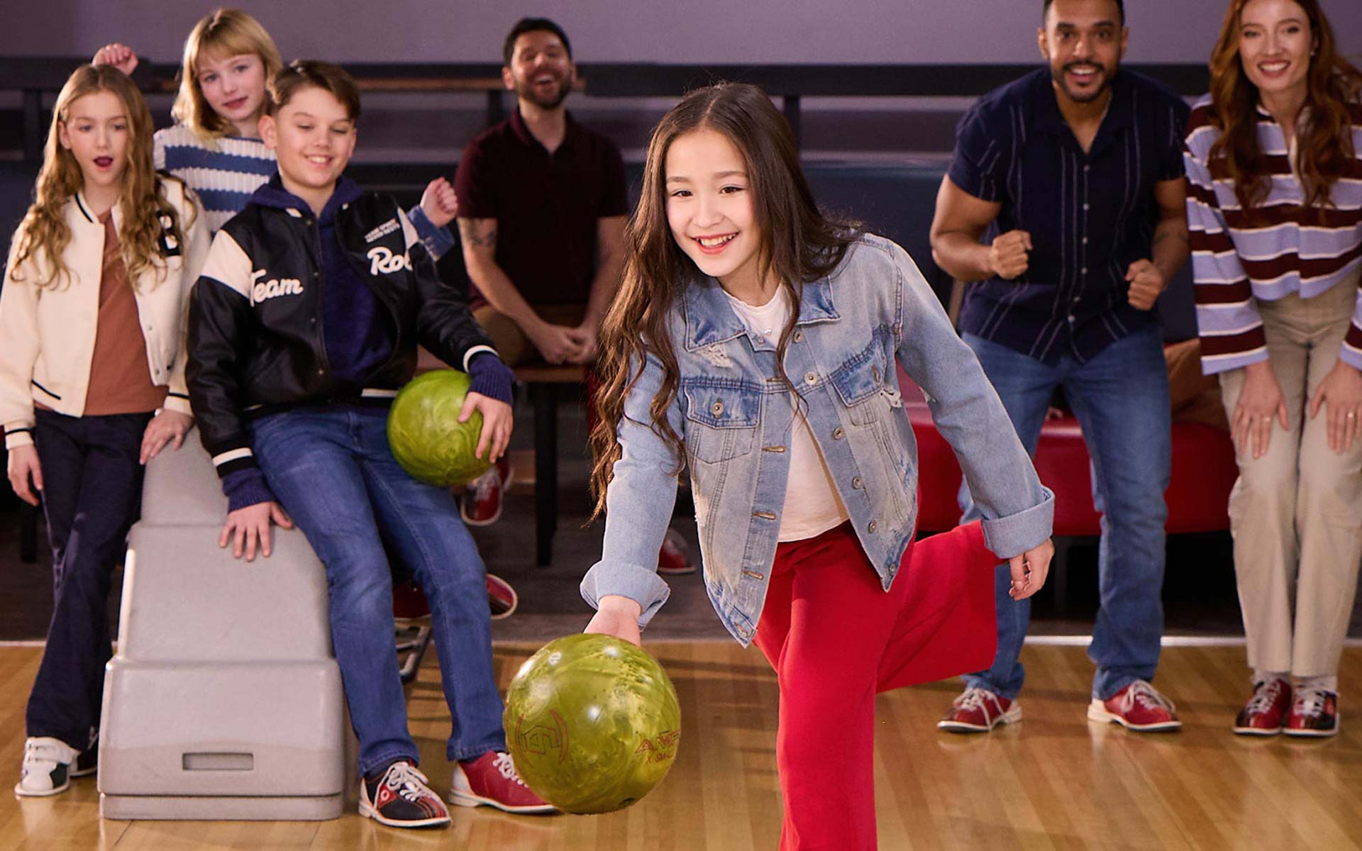 Young girl rolls a bowling ball down the lane while family and friends cheer behind her at an AMF bowling center.