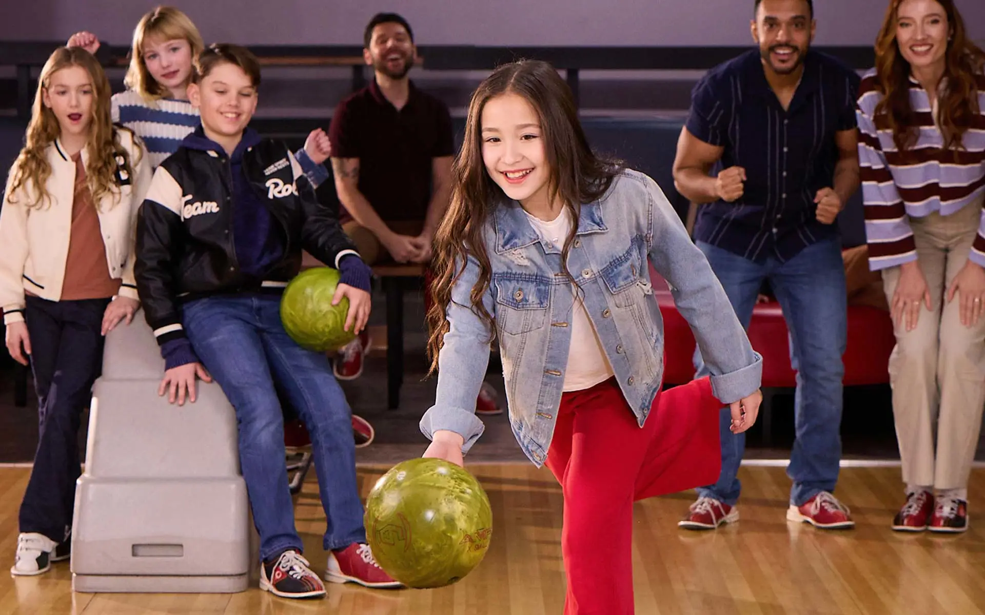 Young girl rolls a bowling ball down the lane while family and friends cheer behind her at an AMF bowling center.