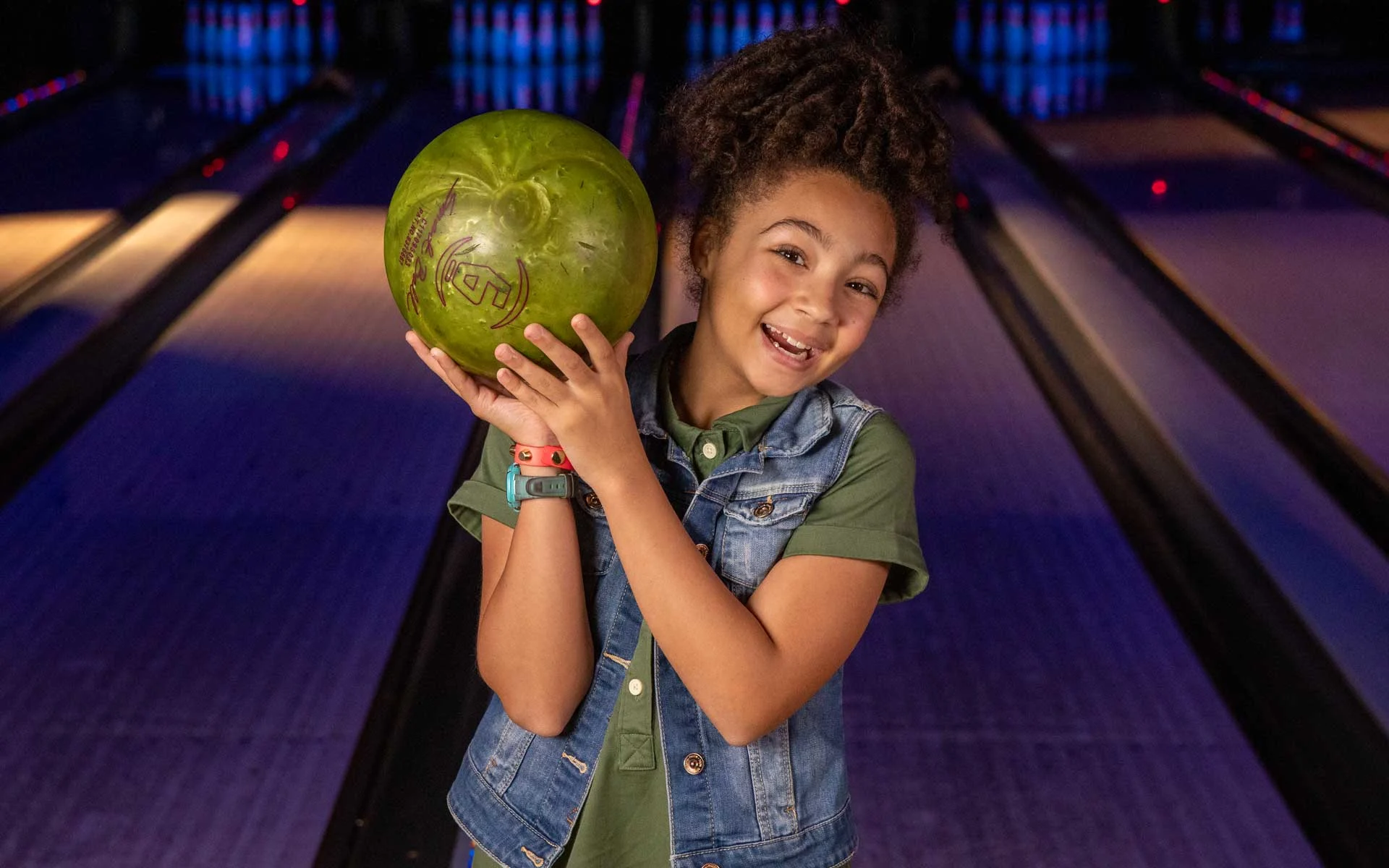 Young girl smiling and holding a green bowling ball on her shoulder while standing on a bowling lane, with glowing pins and a dimly lit alley in the background.