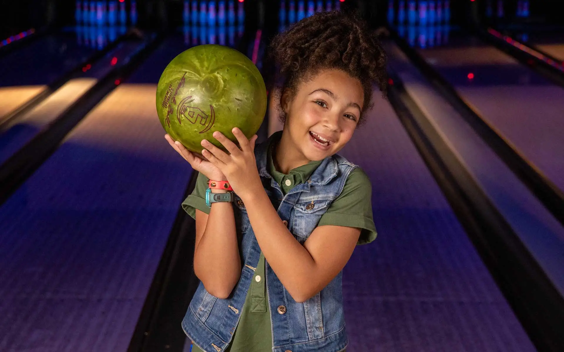 Young girl smiling and holding a green bowling ball on her shoulder while standing on a bowling lane, with glowing pins and a dimly lit alley in the background.