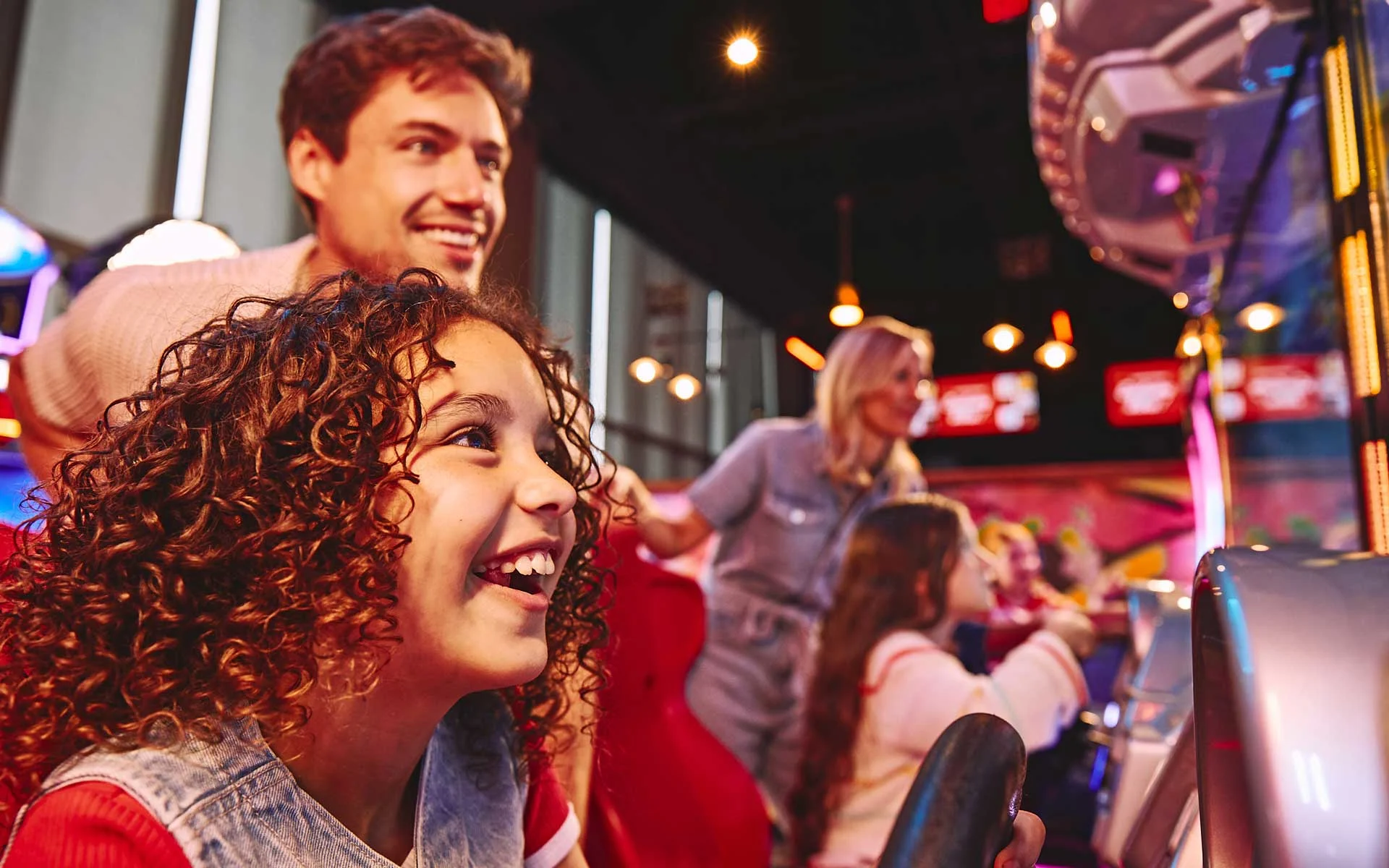 Family enjoying arcade games together at a modern entertainment venue, smiling and engaged in colorful, high-energy gameplay with bright lights and interactive machines.