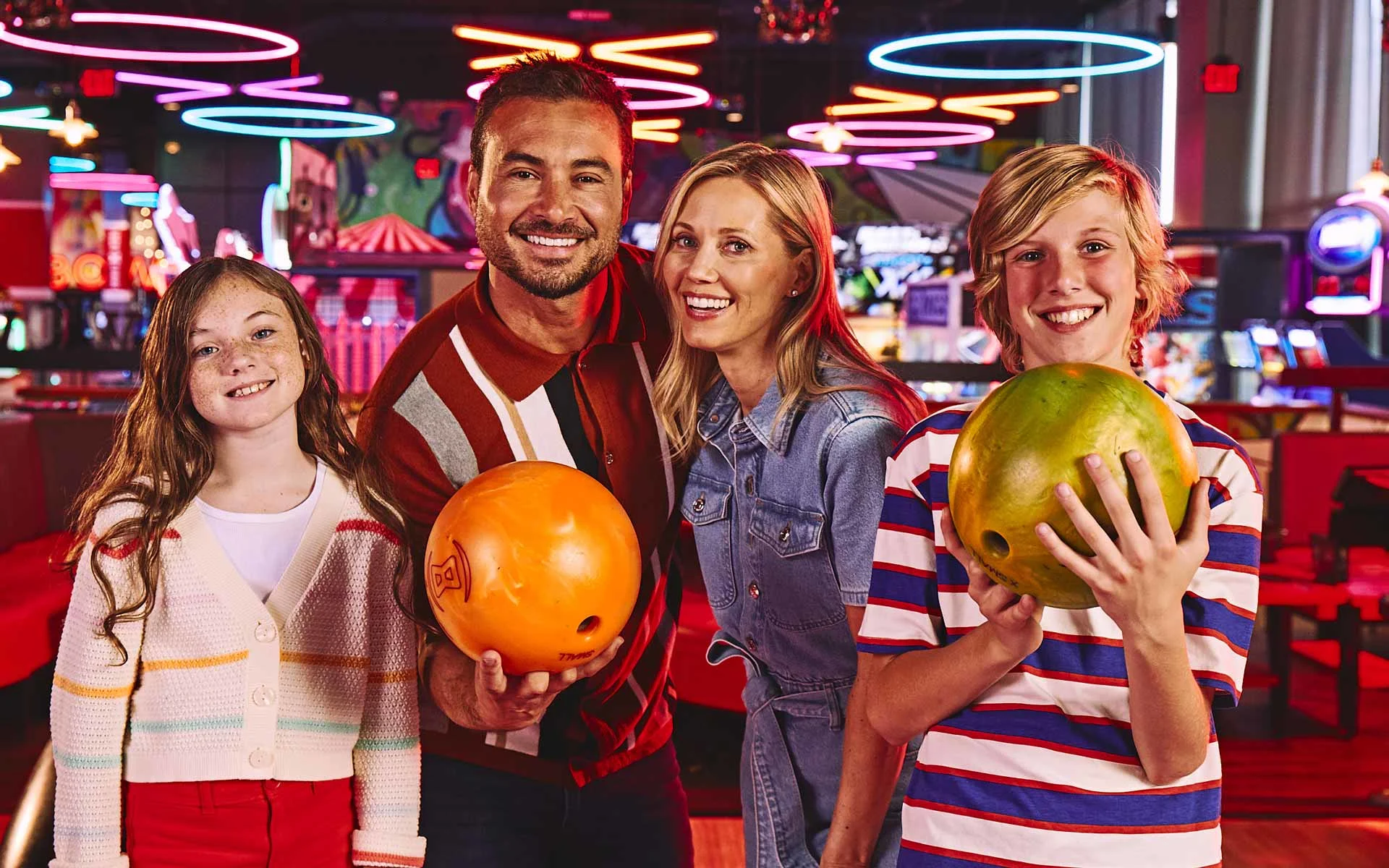 Family smiling and holding bowling balls together in a vibrant bowling center with neon lighting and a fun, welcoming atmosphere