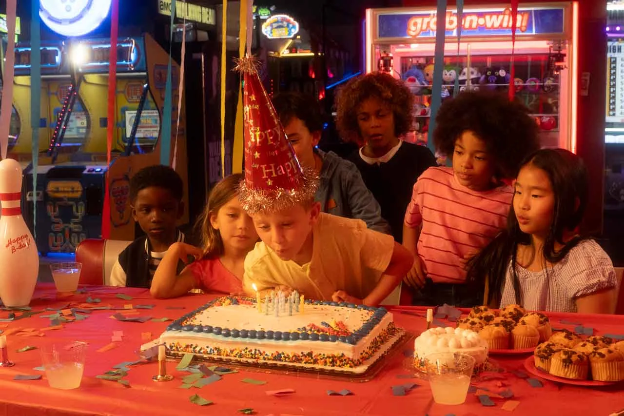 A kids party at an AMF center with a group of kids gathered around a table full of desserts with one child blowing out the cake's candles.
