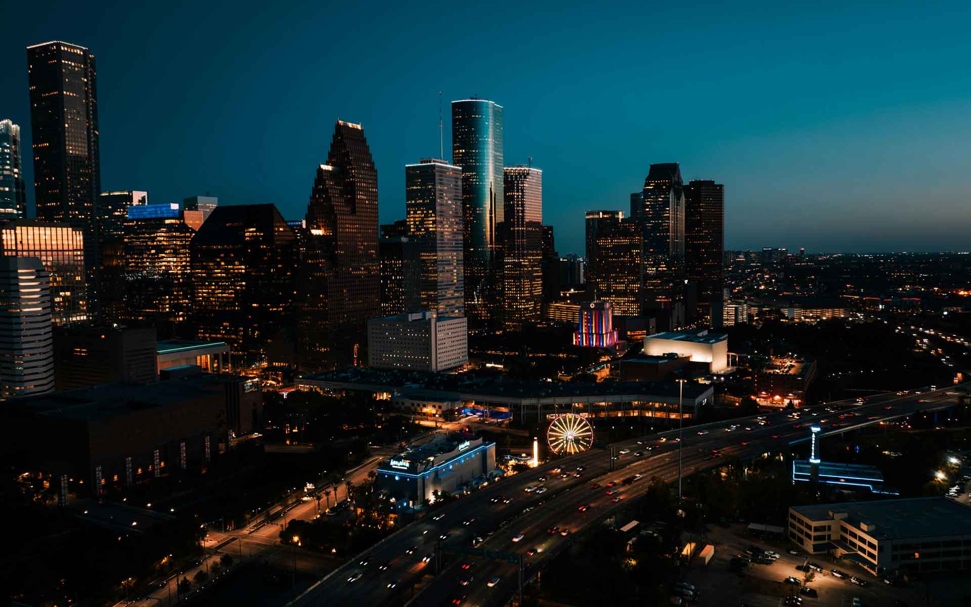 Houston skyline at night with illuminated downtown skyscrapers, highway traffic, and city lights glowing at dusk.