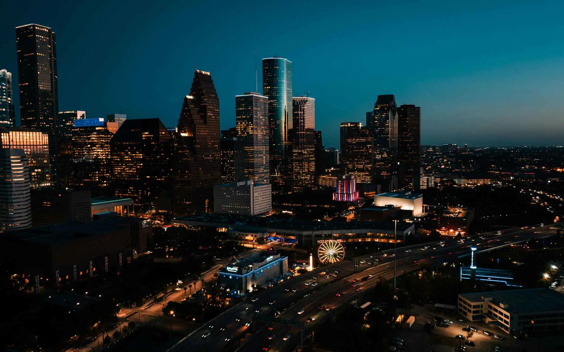 Houston skyline at night with illuminated downtown skyscrapers, highway traffic, and city lights glowing at dusk.