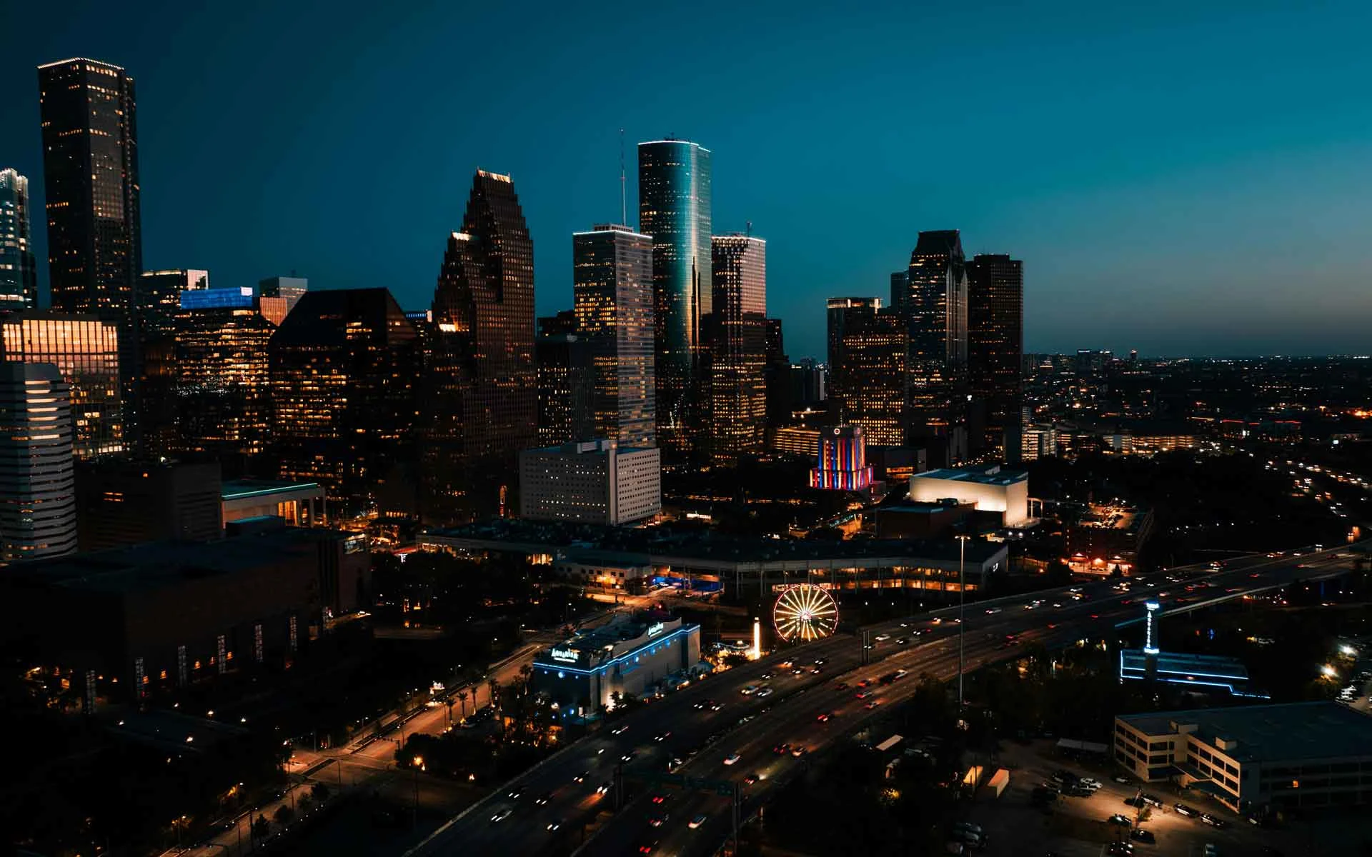 Houston skyline at night with illuminated downtown skyscrapers, highway traffic, and city lights glowing at dusk.